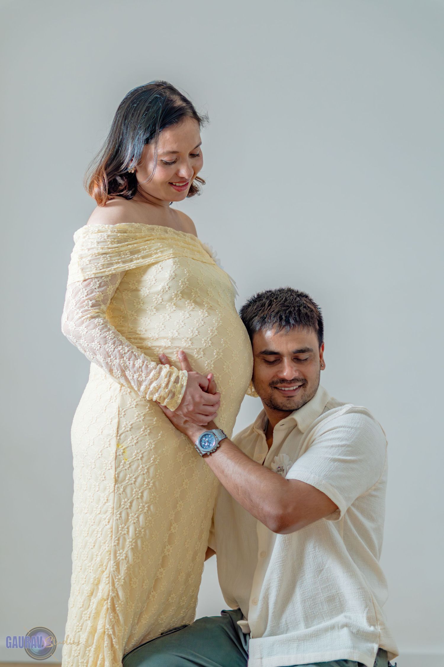 “Pregnant woman and her partner posing together in a studio, smiling and touching the baby bump.”