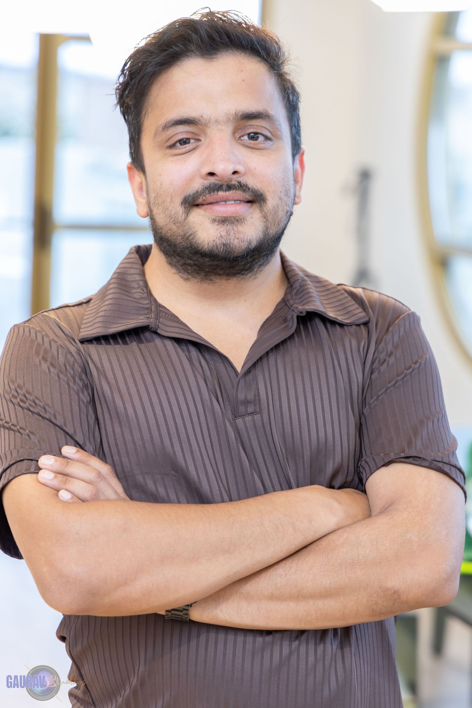 “Portrait of a man posing indoors, standing casually with hands folded together.”