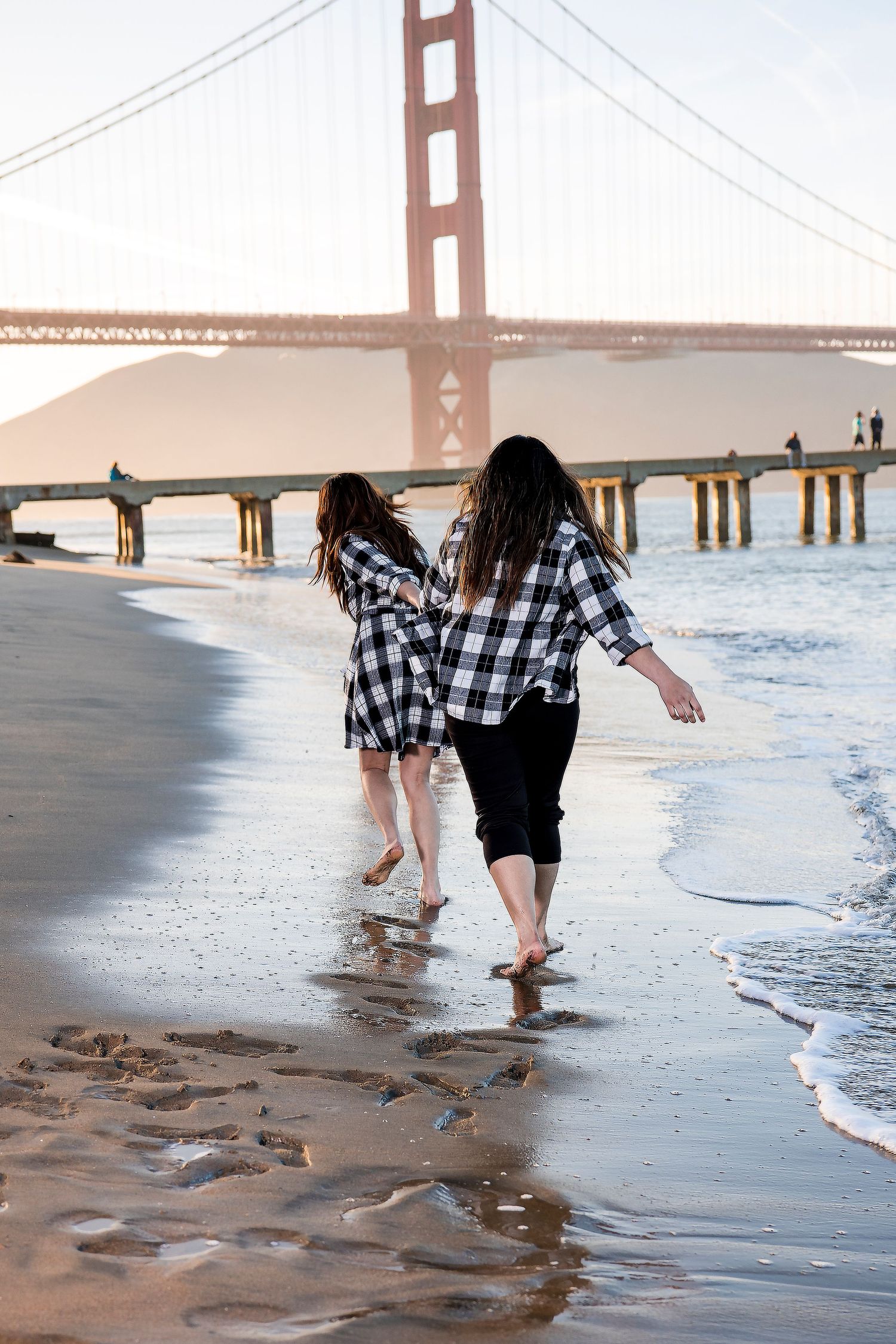 Couple playfully splashing in waves at beach with Golden Gate Bridge visible in background.