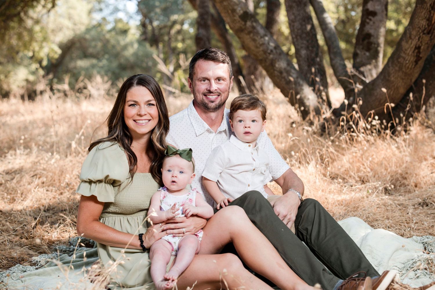 Family sitting together on a blanket in a sunny outdoor setting with trees in the background.