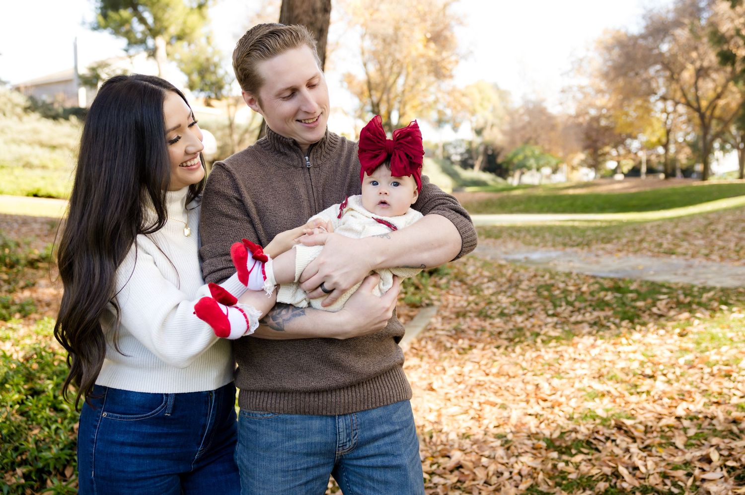 A couple sits together on fallen autumn leaves in a park with their baby during a fall photo session.