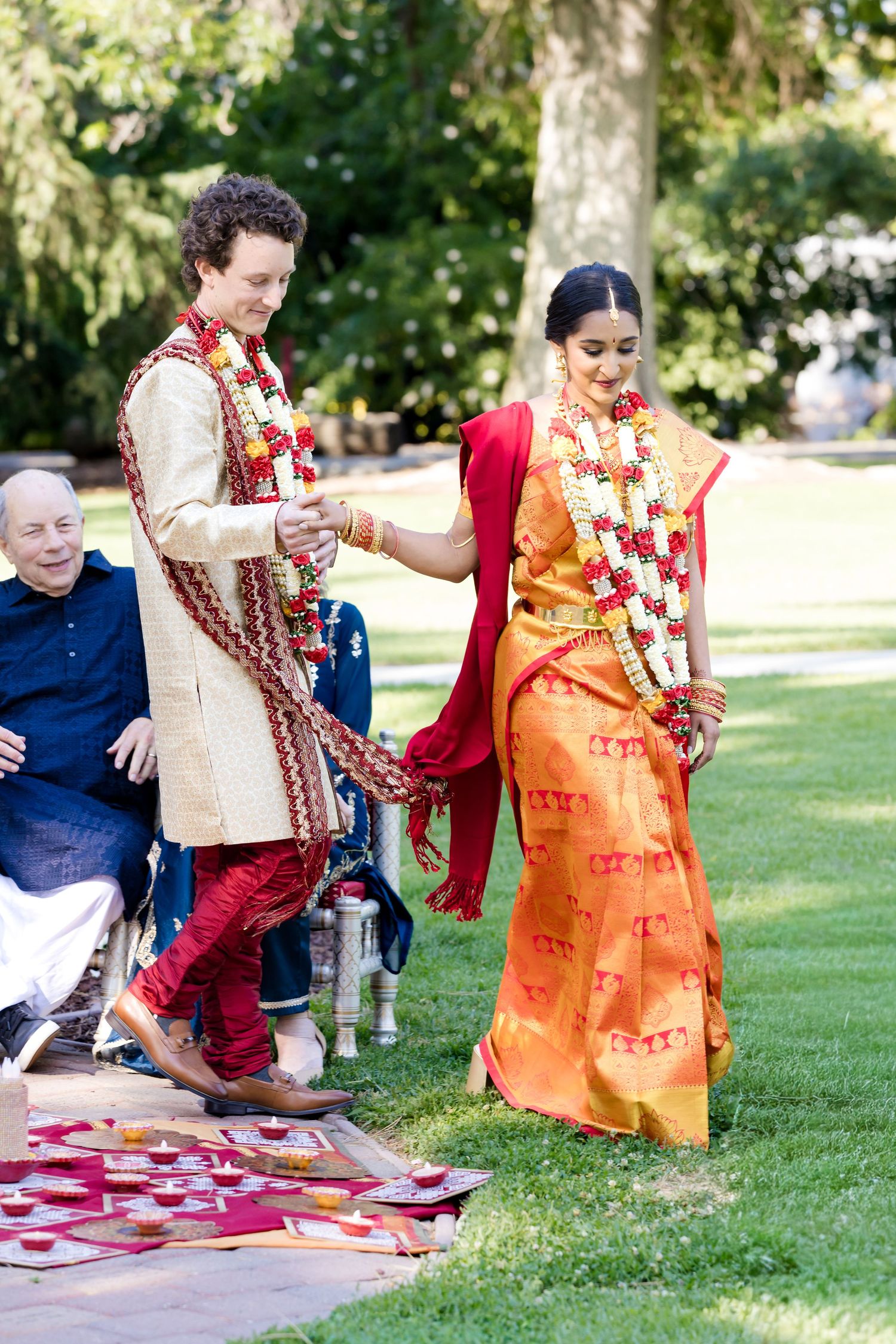 Couple in traditional Indian wedding attire plays games on red and white checkered blanket outdoors.