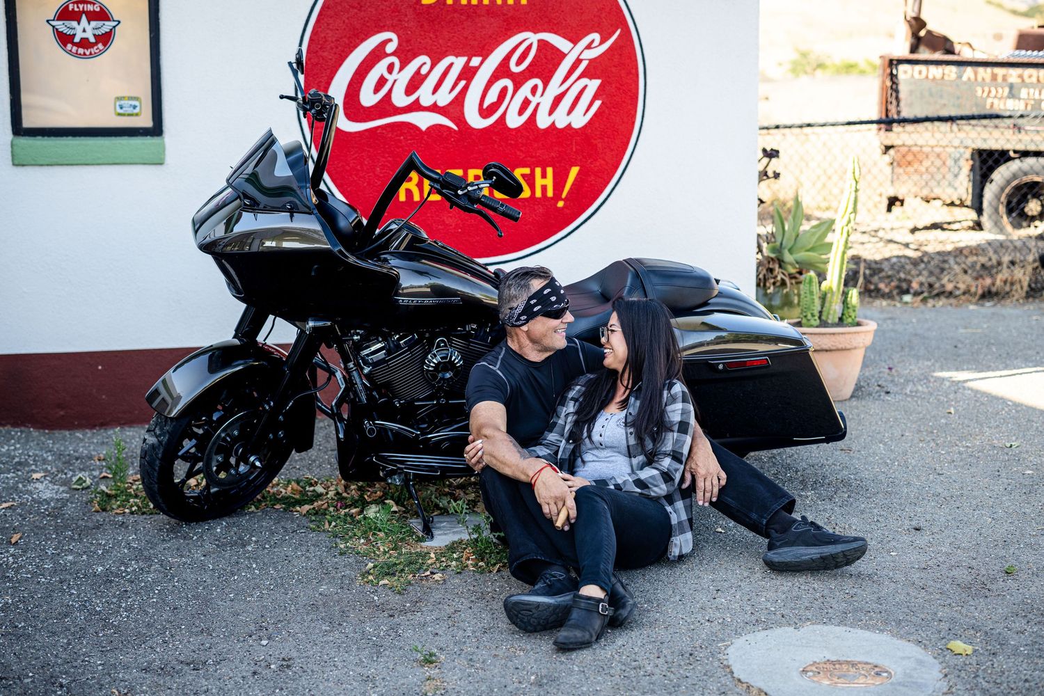 A motorcycle rider rests against a black touring bike parked beneath a vintage Coca-Cola sign.