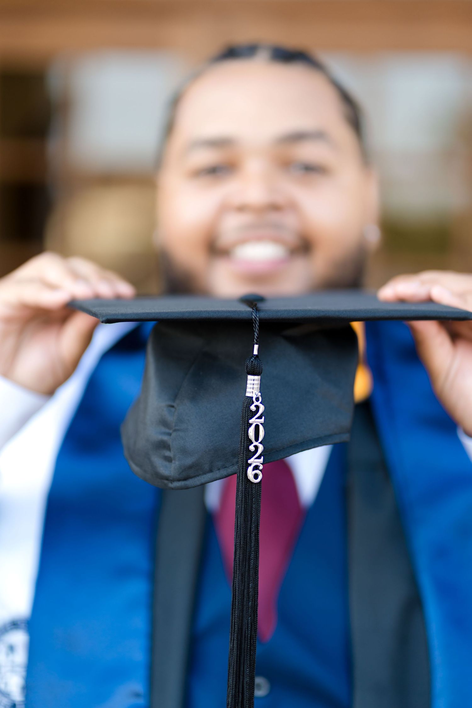 Smiling graduate in blue gown holds up black 2026 graduation cap toward camera in celebratory portrait photo.