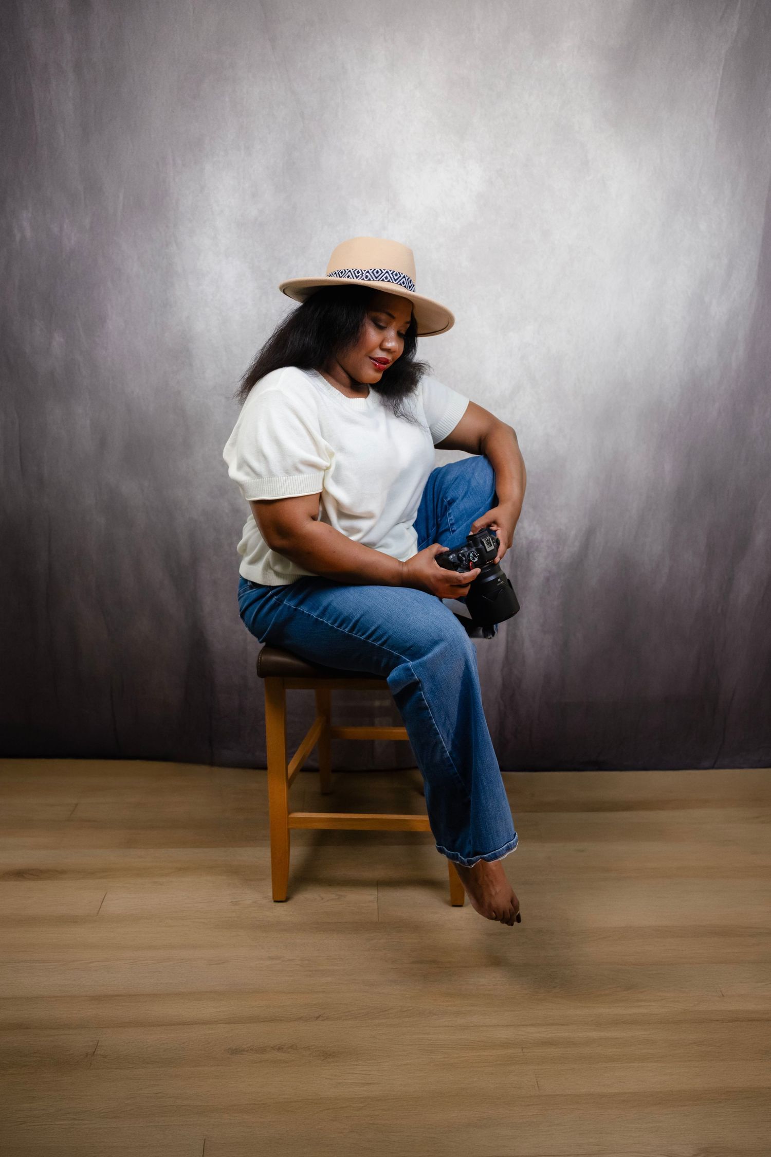 Woman in white tee, jeans, and straw hat sits on wooden stool holding camera in studio setting.