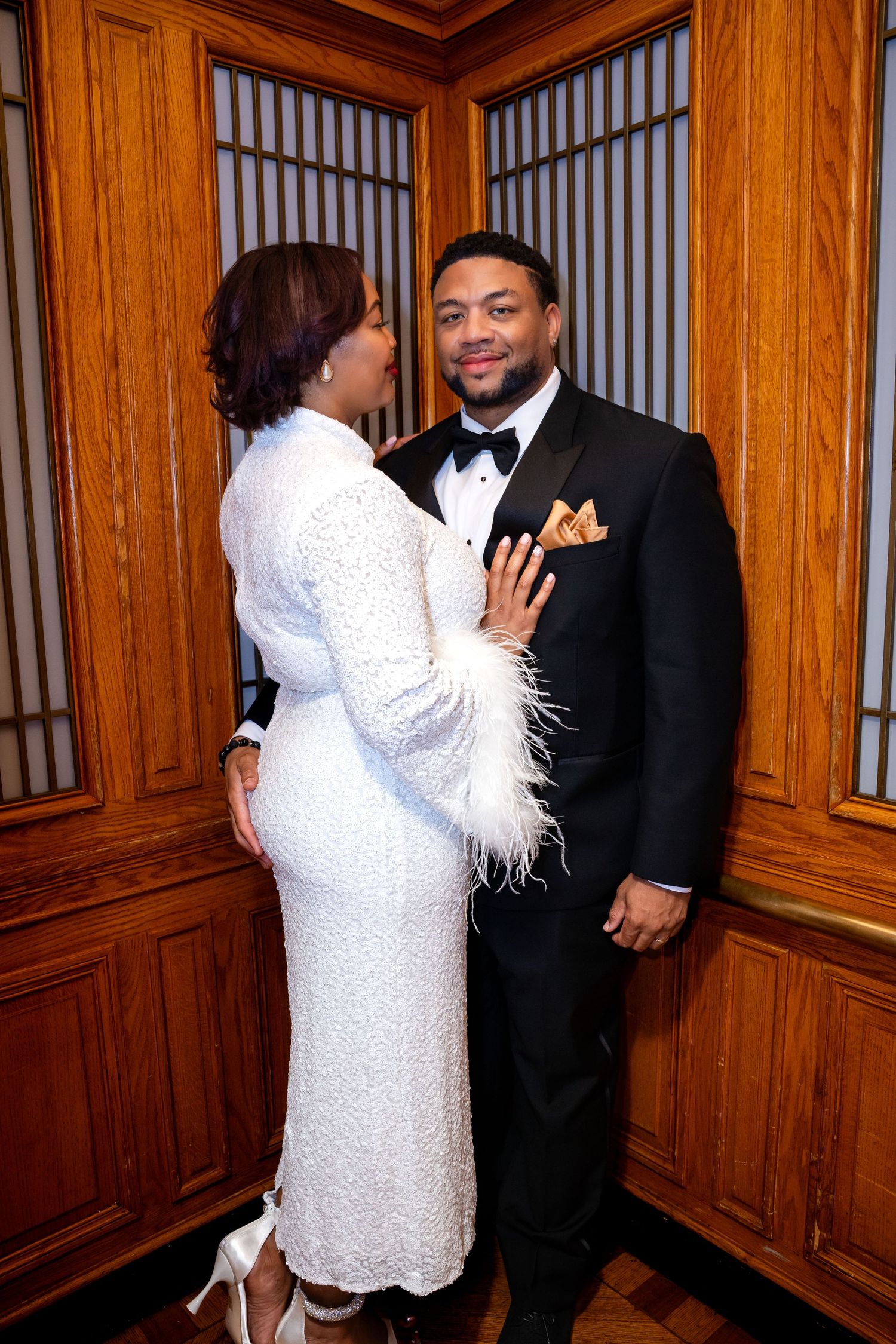 Formal couple in elegant white dress and black tuxedo posing in wood-paneled room.