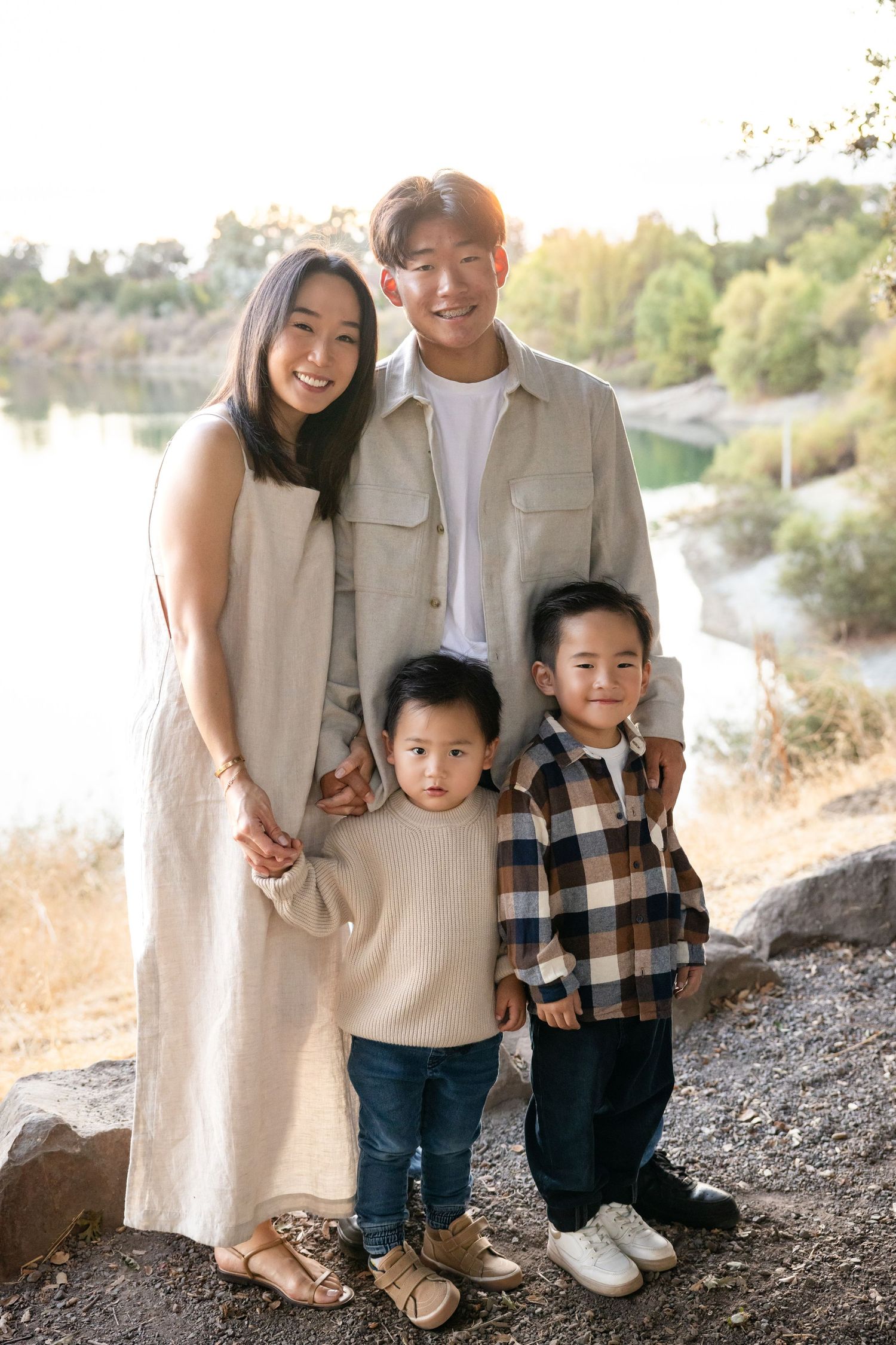 A family of four poses together outdoors on a hillside path during golden hour in neutral-colored clothing.