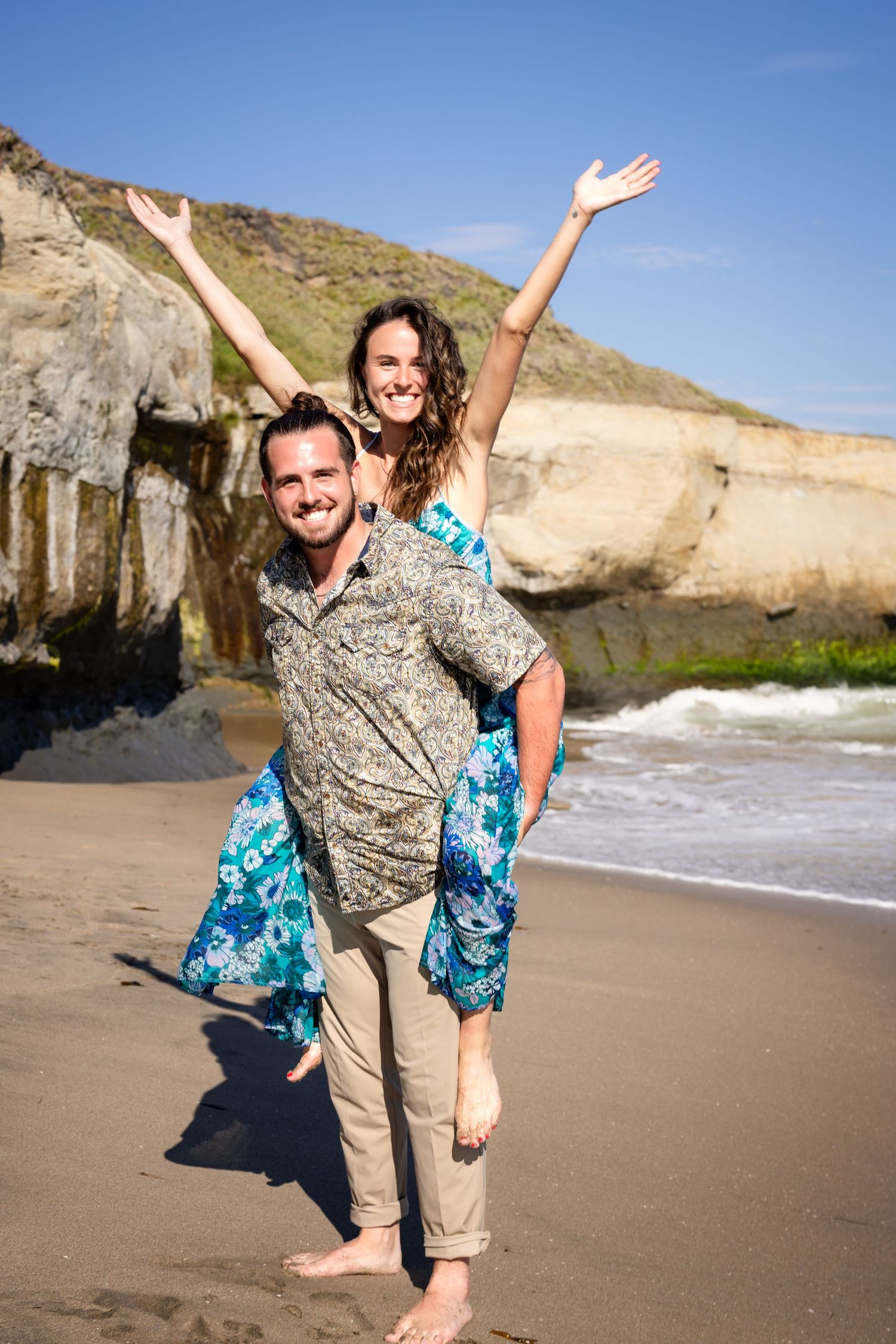 Couple playing and posing playfully on sandy beach with cliffs in background.