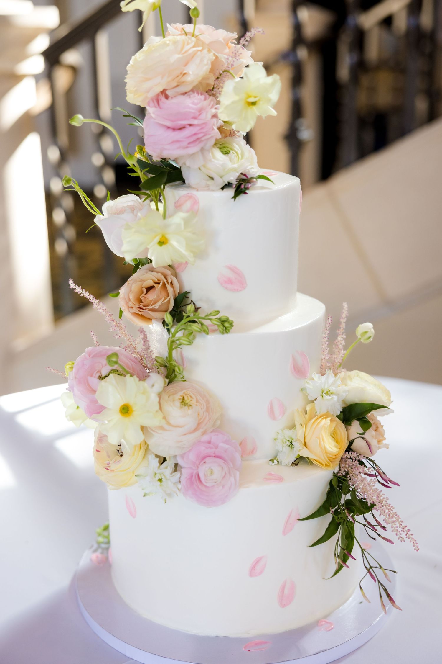 Elegant white wedding cake decorated with delicate pink and peach roses and greenery on a light-filled table setting.