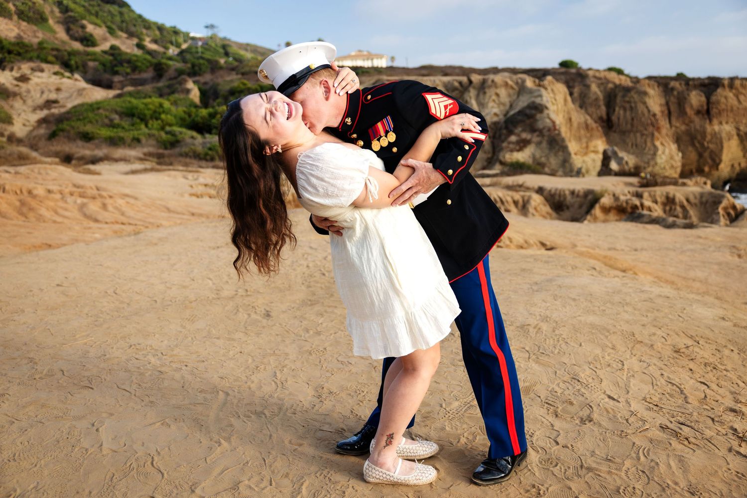 Marine in dress uniform dips and kisses woman in white dress on sandy beach with rocky cliffs.