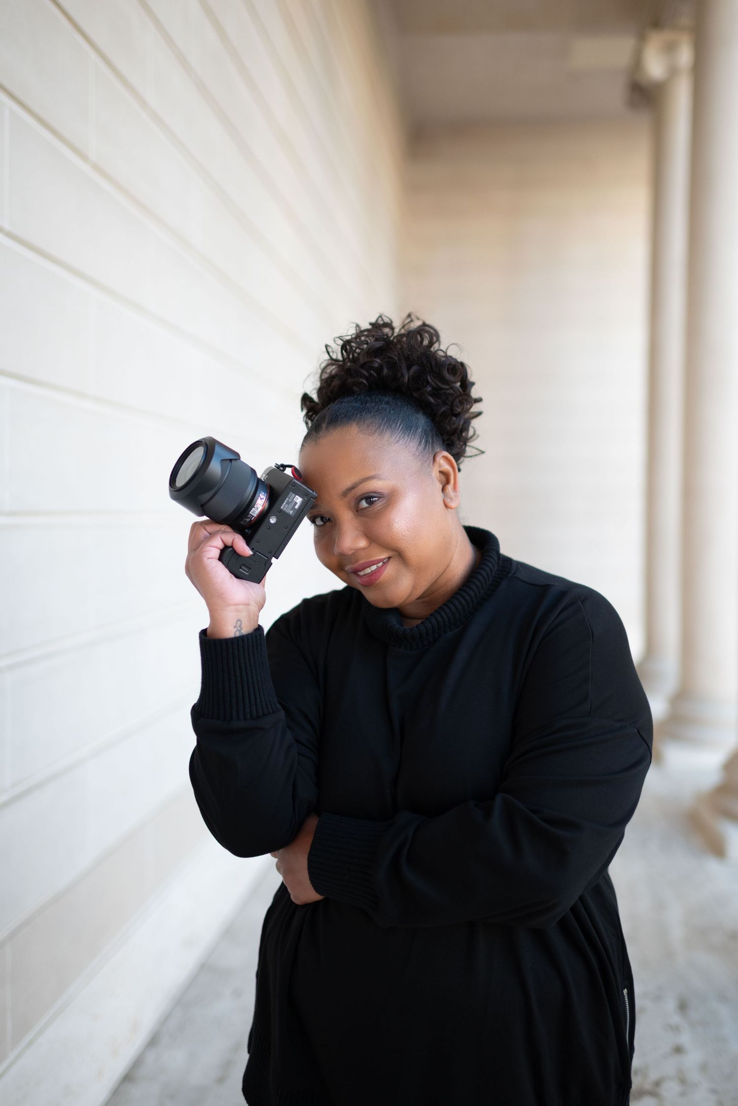 Professional photographer in black clothing holding camera and posing against white wall.
