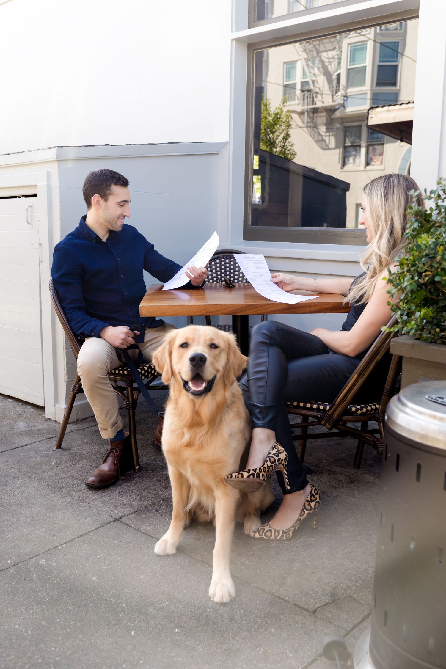 A Golden Retriever sits attentively next to people reviewing documents at an outdoor cafe table.
