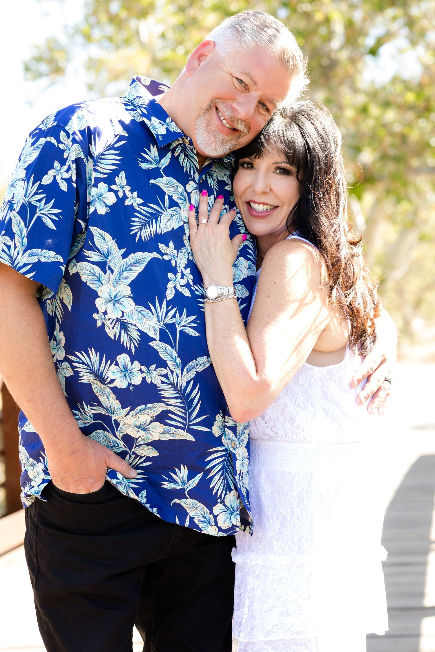 Couple embraces outdoors wearing coordinating blue Hawaiian shirt and white lace dress in bright sunlight.