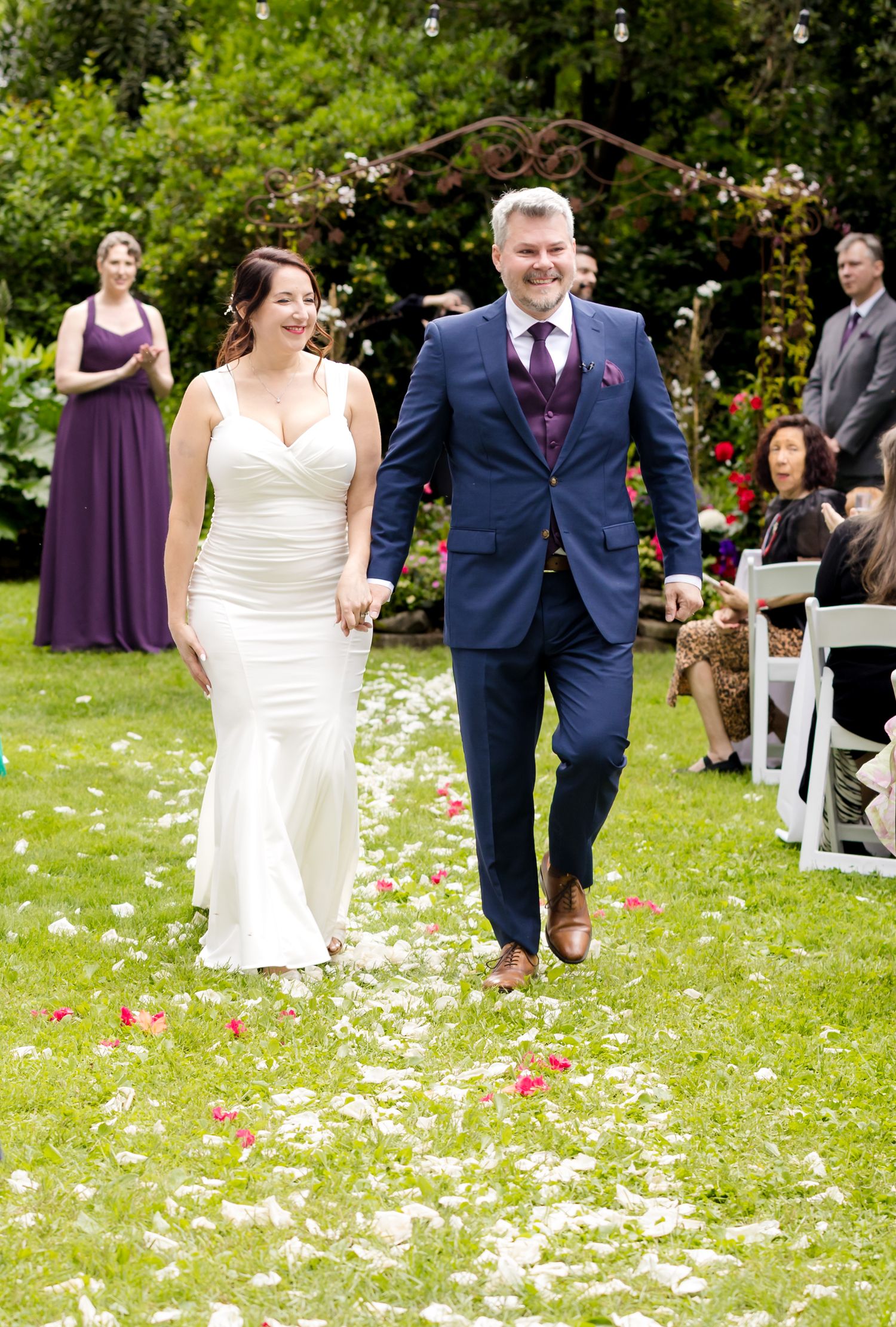 Couple walks down an aisle covered in rose petals at an outdoor garden wedding ceremony.