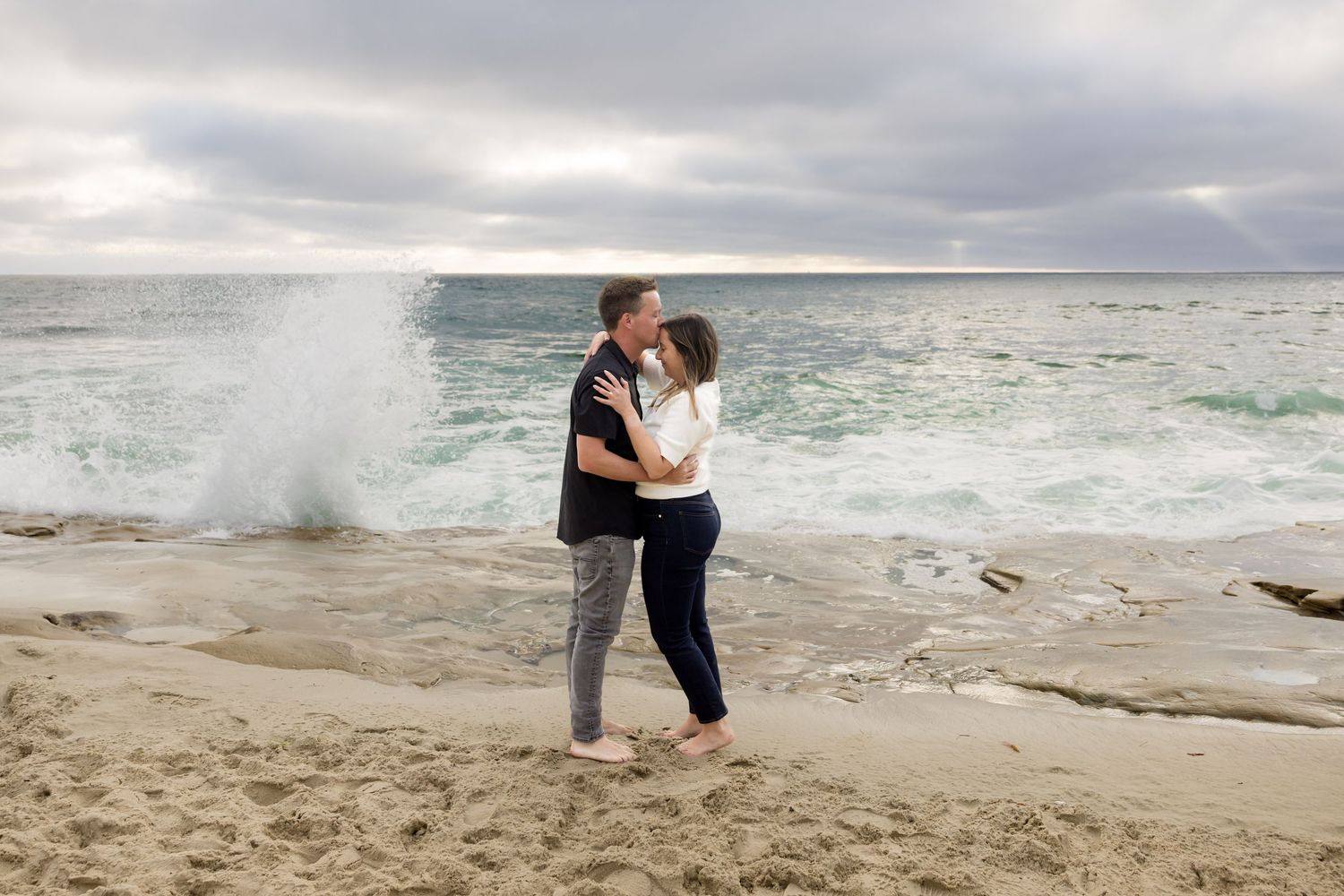 Couple kissing on a sandy beach with dramatic ocean waves crashing behind them under a cloudy sky.