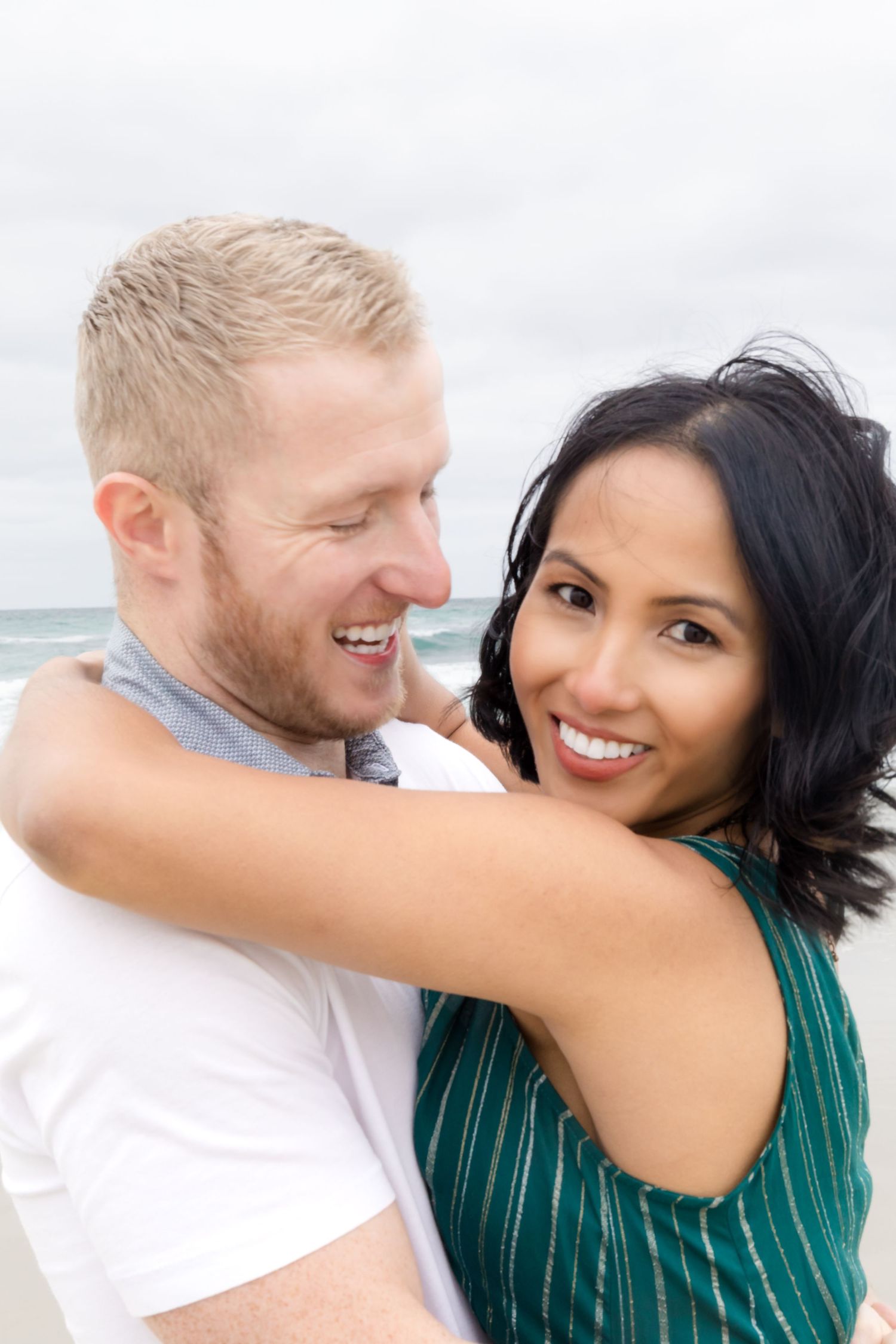 Couple embraces on beach with bright smiles in casual green and white outfits.