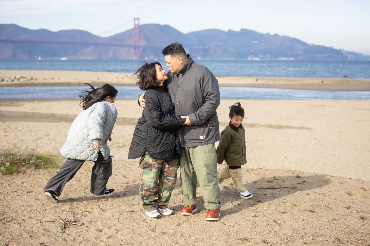 Family members playing and interacting on sandy beach with bridge in background.