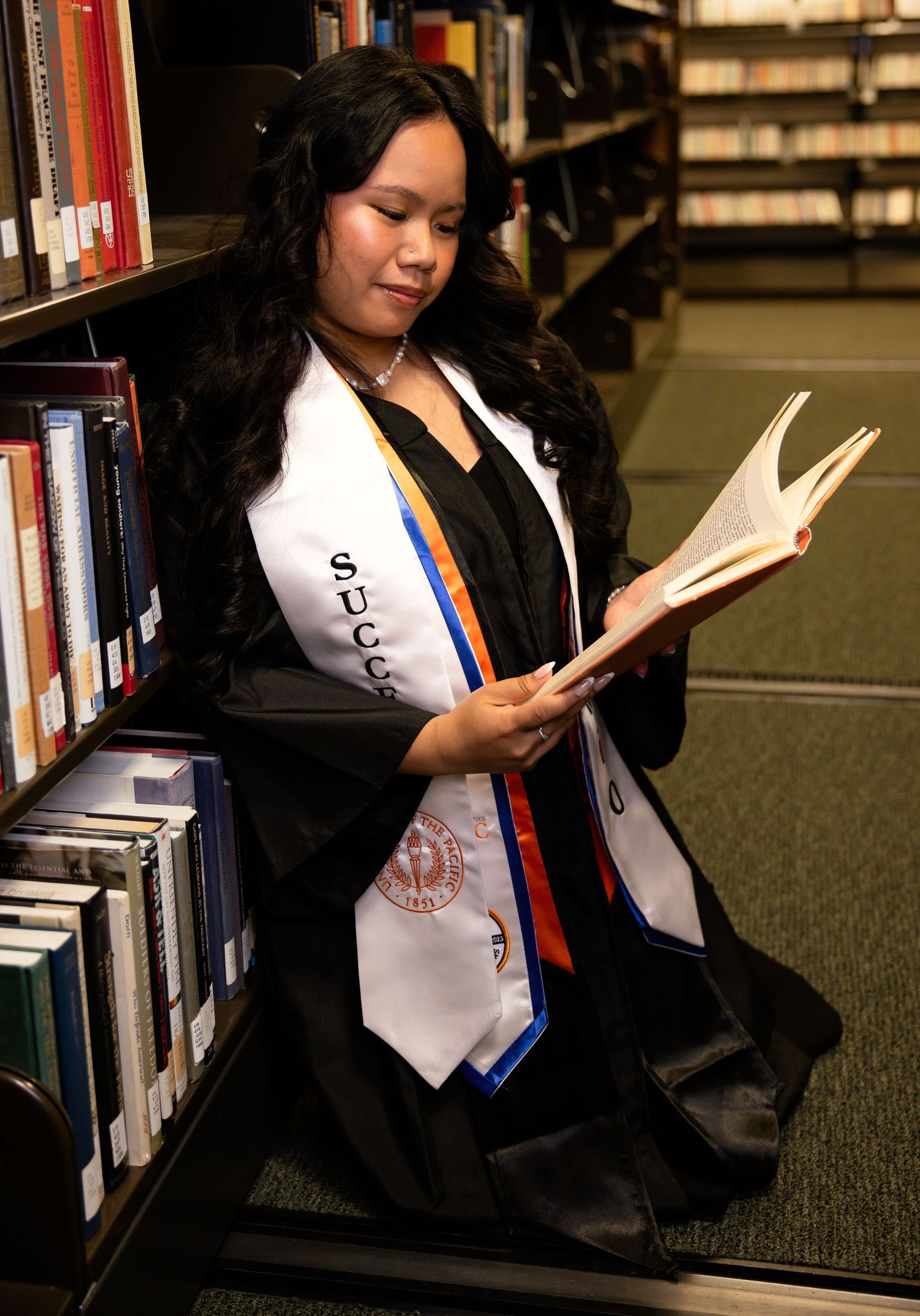 Student reading in library stacks while wearing graduation attire.
