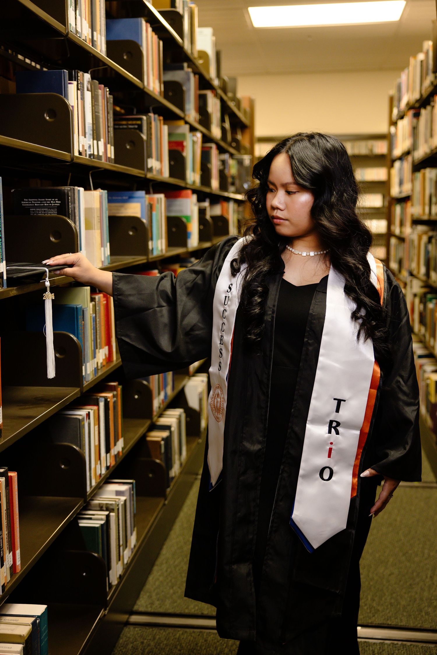 A person wearing graduation attire browses through library bookshelves in a dimly lit academic setting.