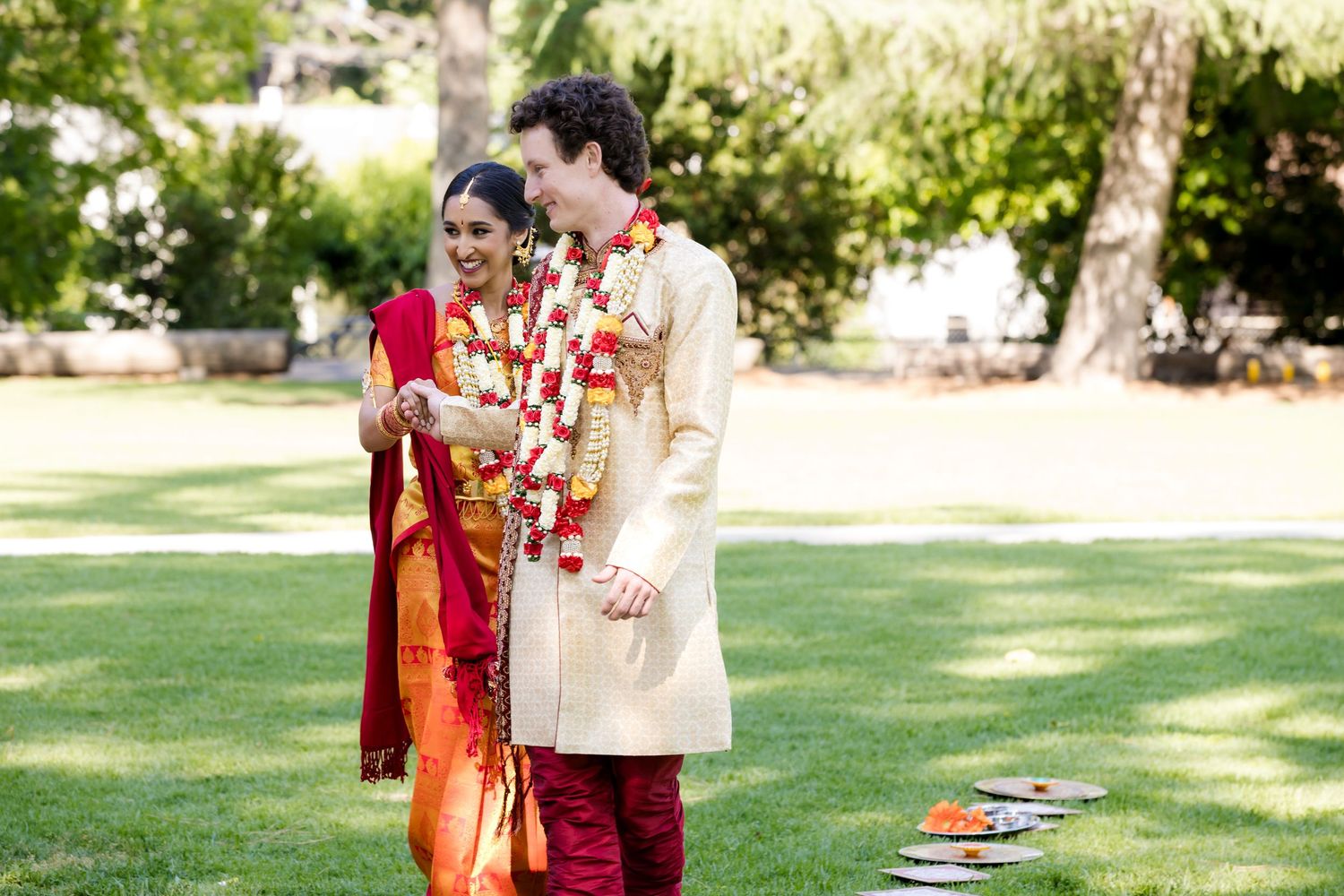 A couple in traditional Indian wedding attire share a tender moment on a sunlit lawn during their celebration.