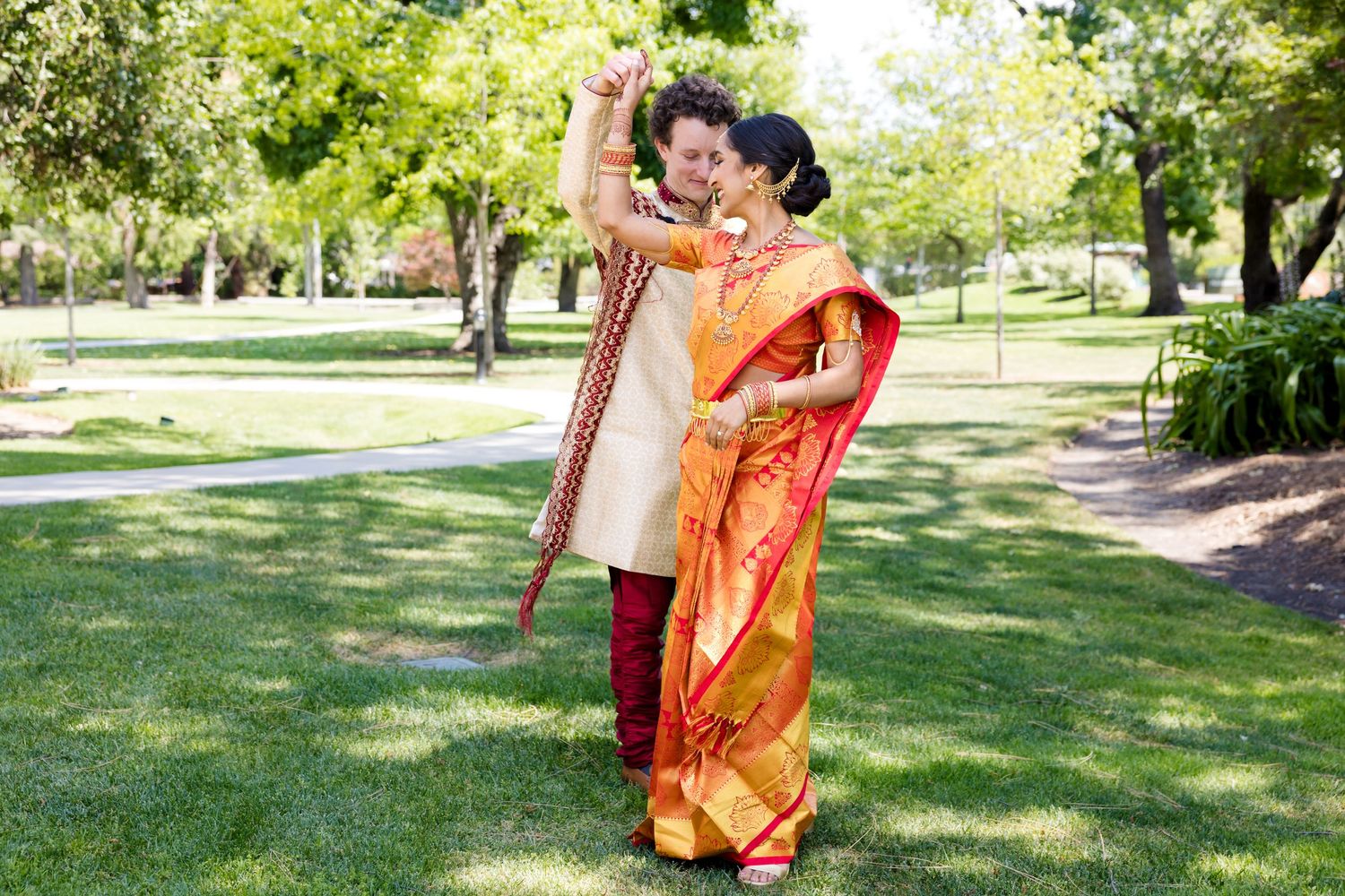 Couple in traditional Indian wedding attire embracing in sunny garden setting.