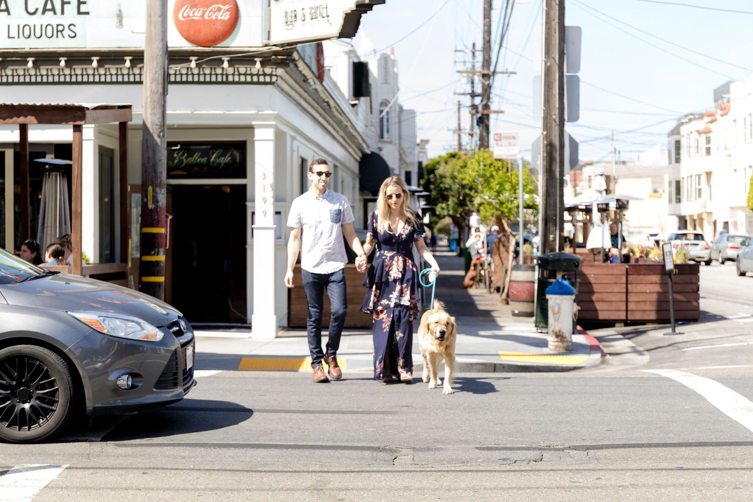 Family walking their dog on a sunny urban street corner near a cafe.