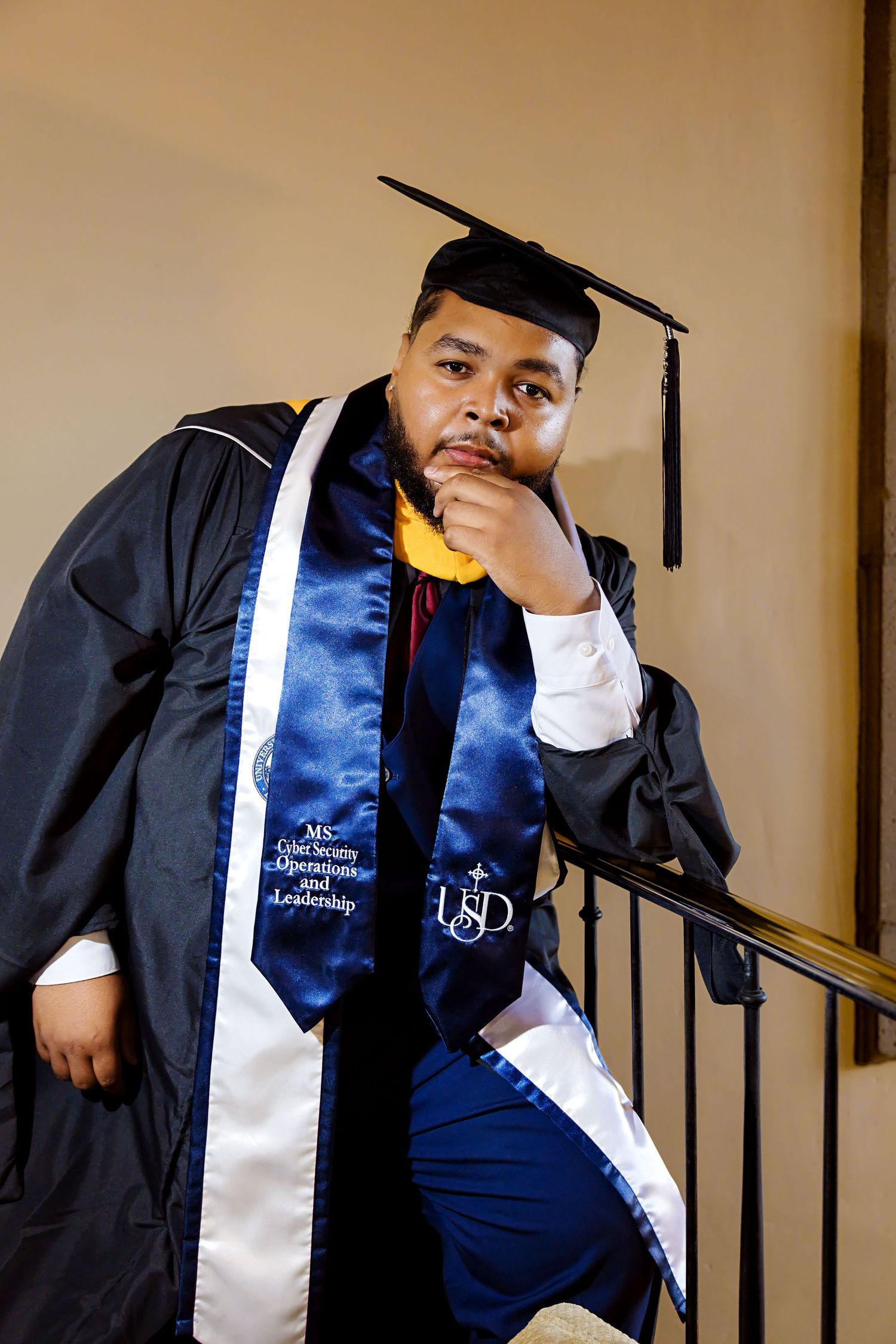 Graduate in black cap and gown with USD blue sash poses thoughtfully on staircase railing.