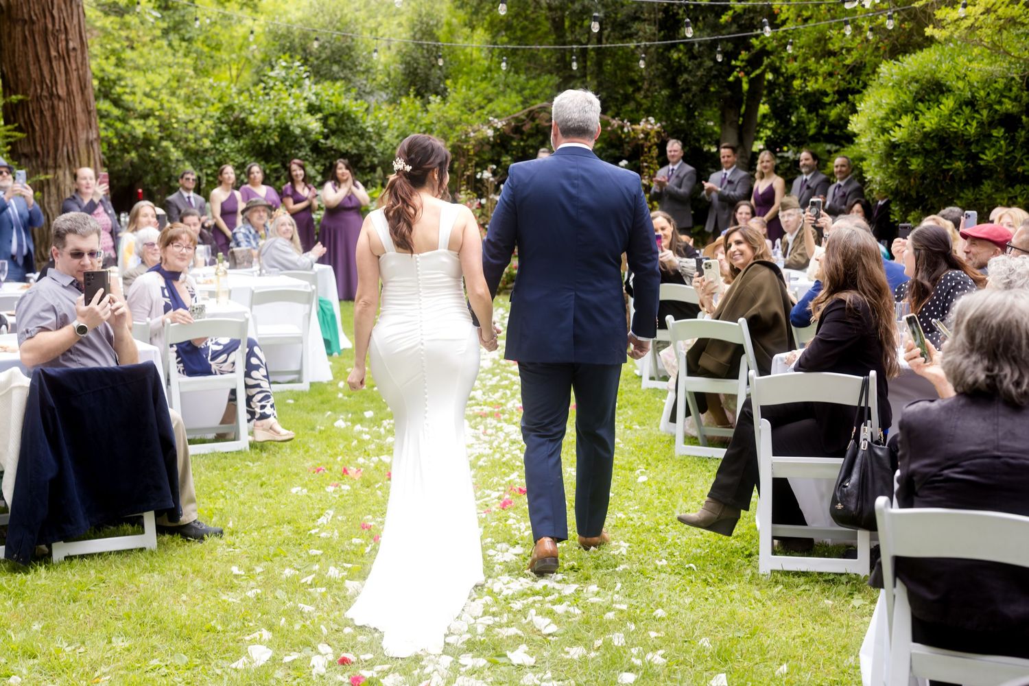 Wedding guests mingle outdoors at a garden reception with white chairs and lush greenery.