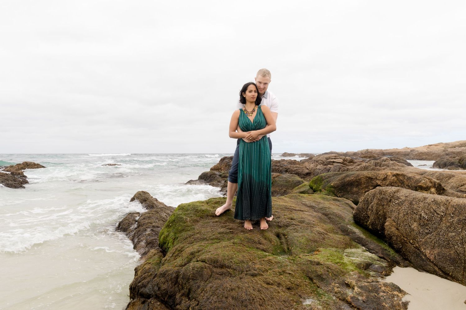 Woman in teal dress poses on rocky coastal overlook with ocean waves in background.