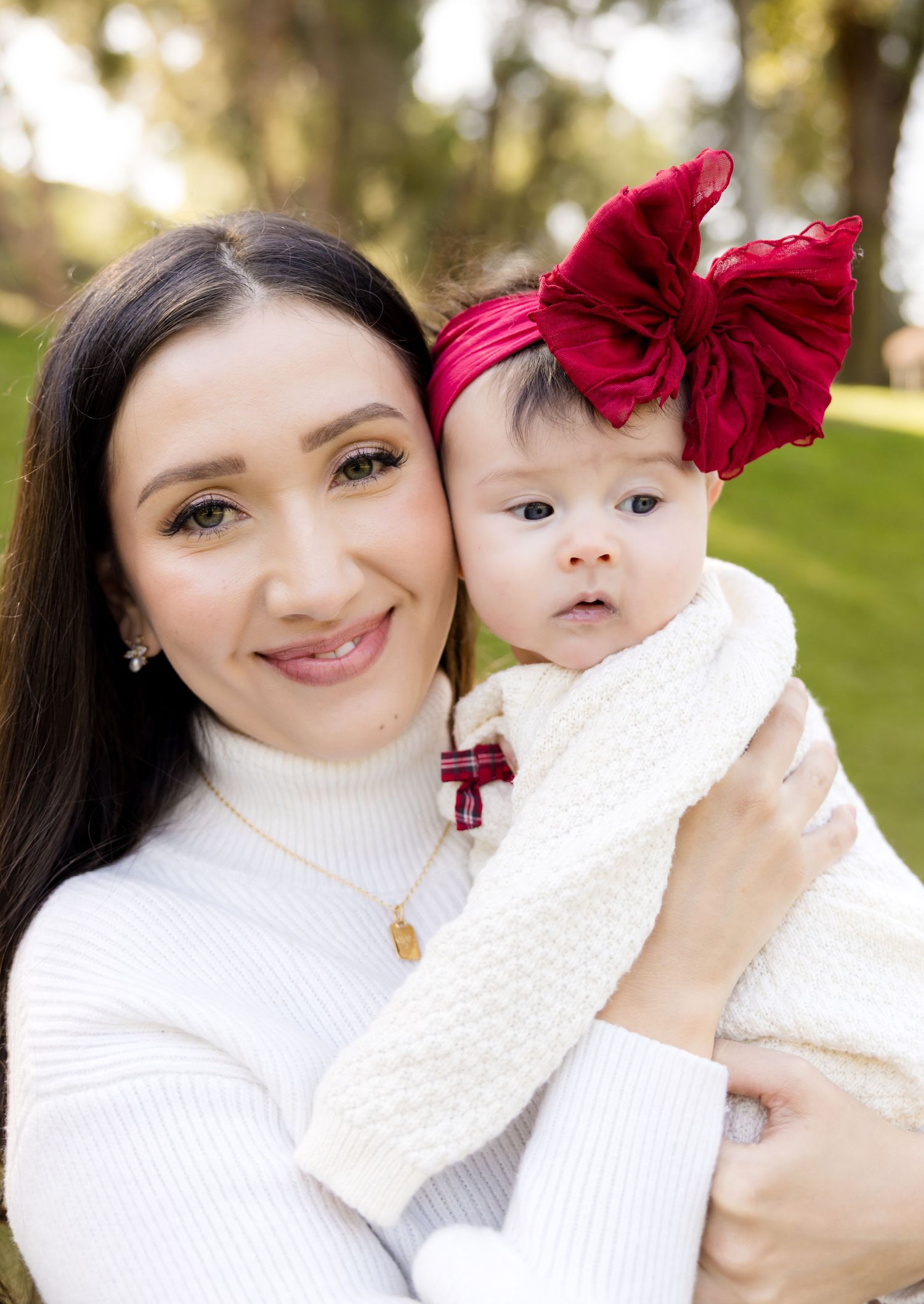 A mother in a white sweater holds a baby wearing a large red bow headband outdoors in a park setting.