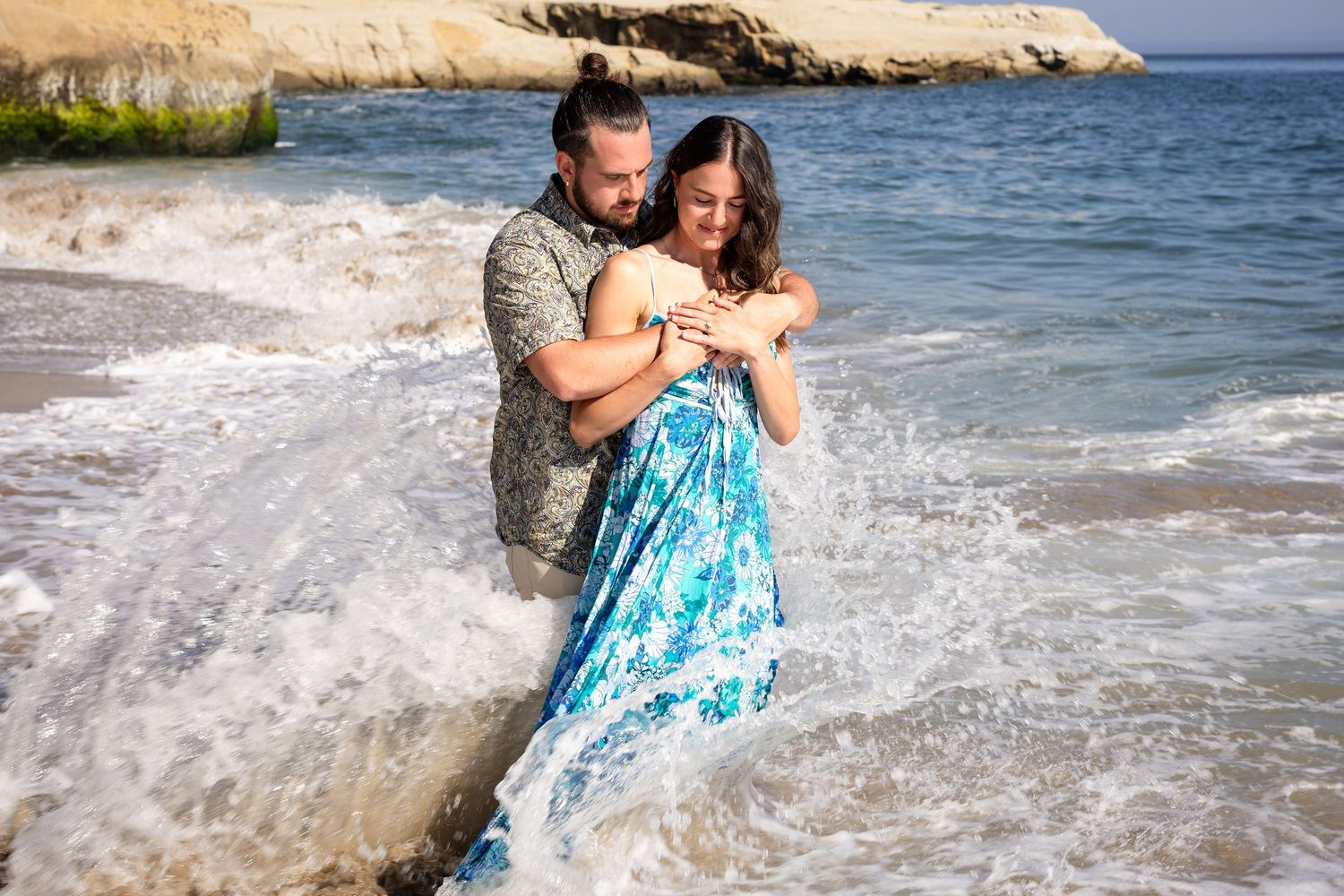 Couple embracing on rocky beach with ocean waves splashing around them.