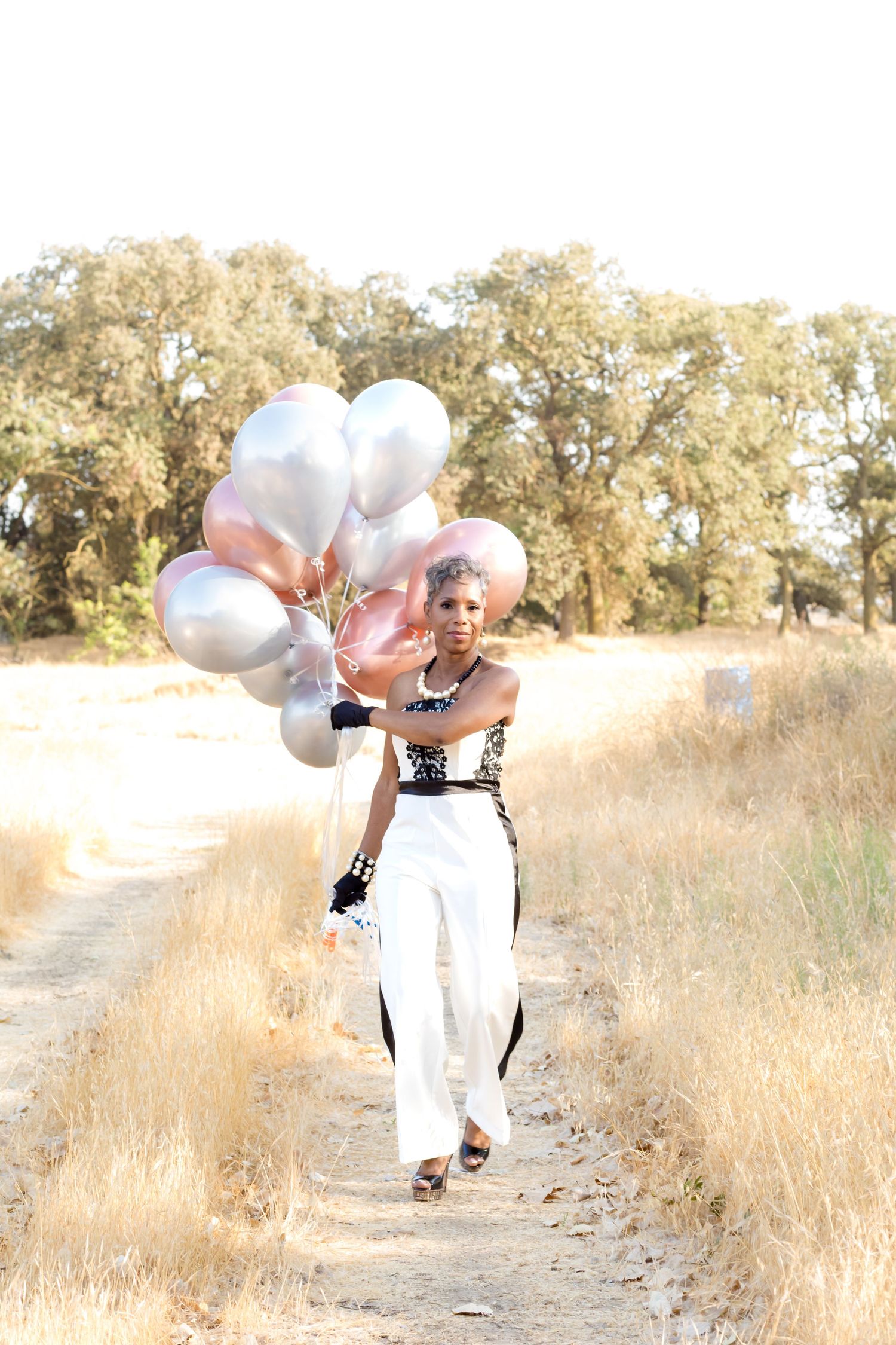 A person in white clothing runs down a dirt path holding white balloons in a golden field on a sunny day.