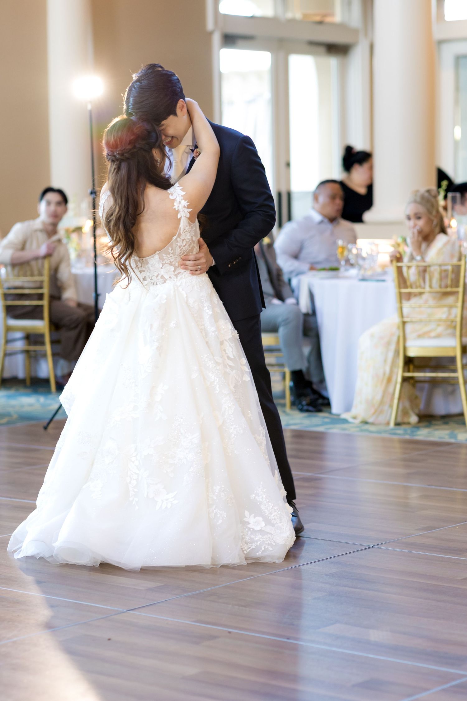 Newlyweds share their first dance in an elegant reception hall with guests seated at tables.
