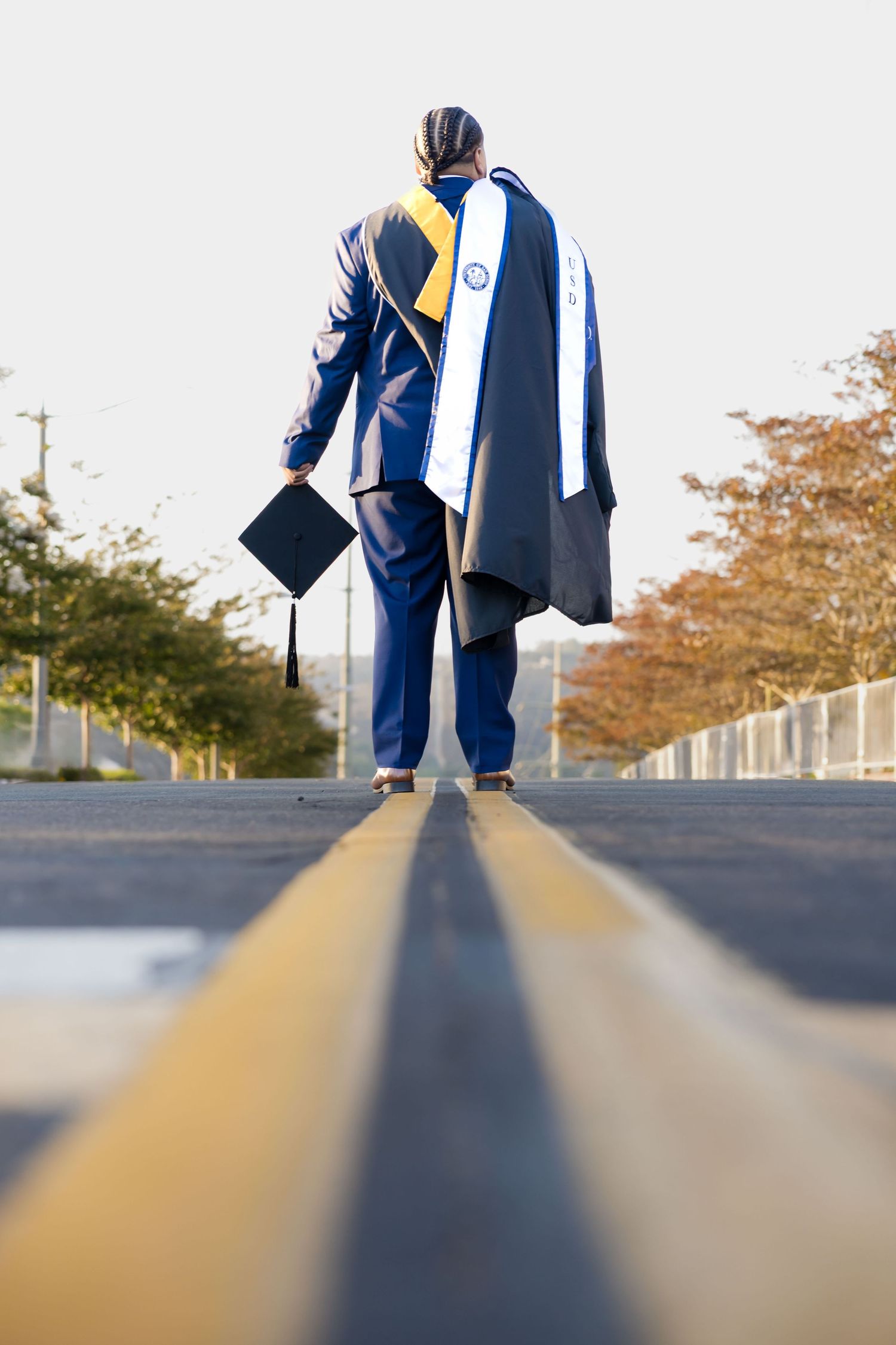 Graduate in cap and gown walks down empty road holding mortarboard, viewed from behind on graduation day.