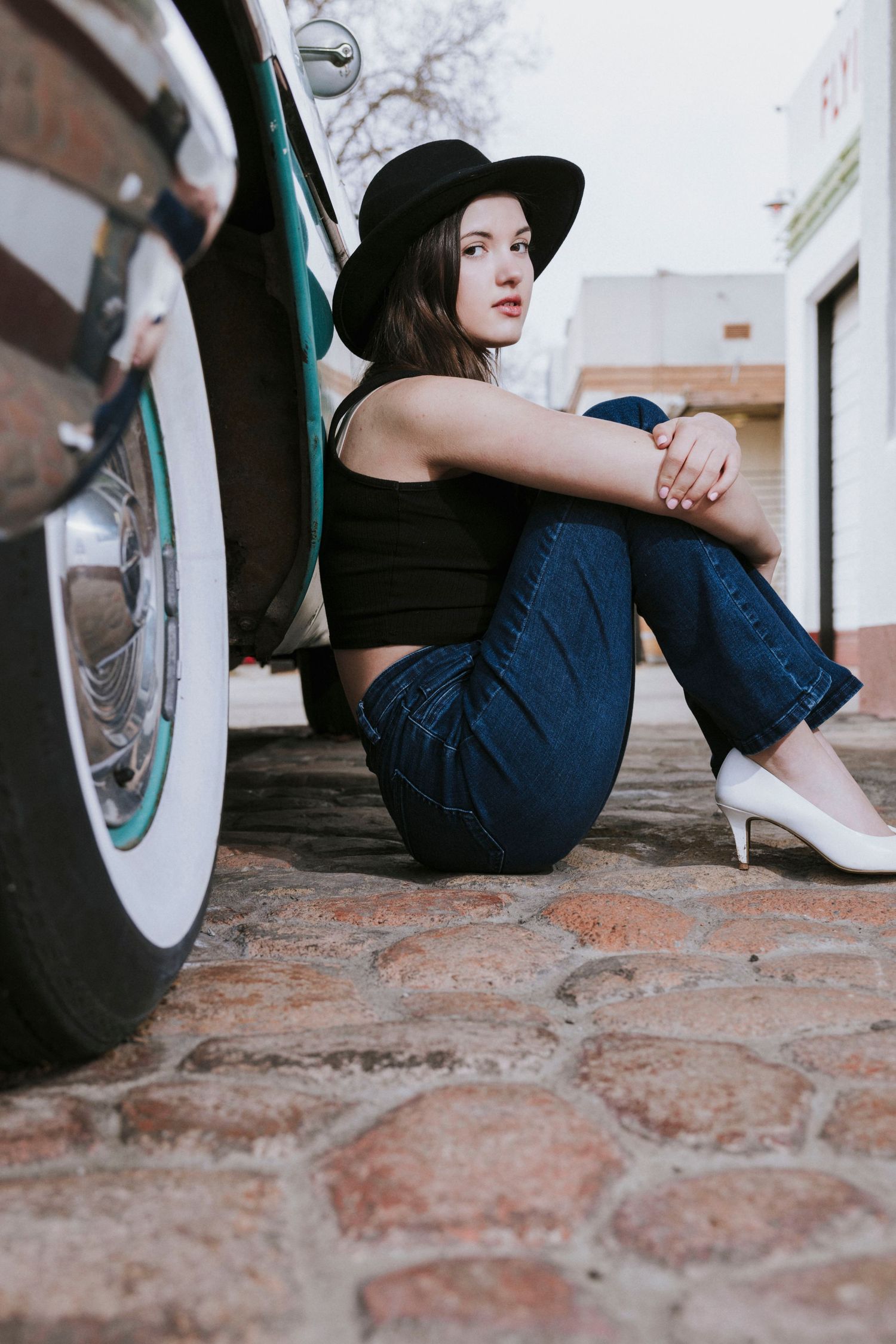 Person in black outfit and hat sitting against white vehicle on brick ground.