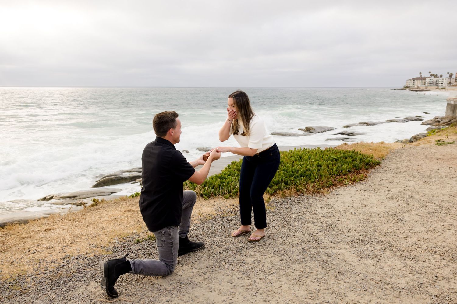 Man proposes on one knee to surprised woman by rocky ocean shore on overcast day.