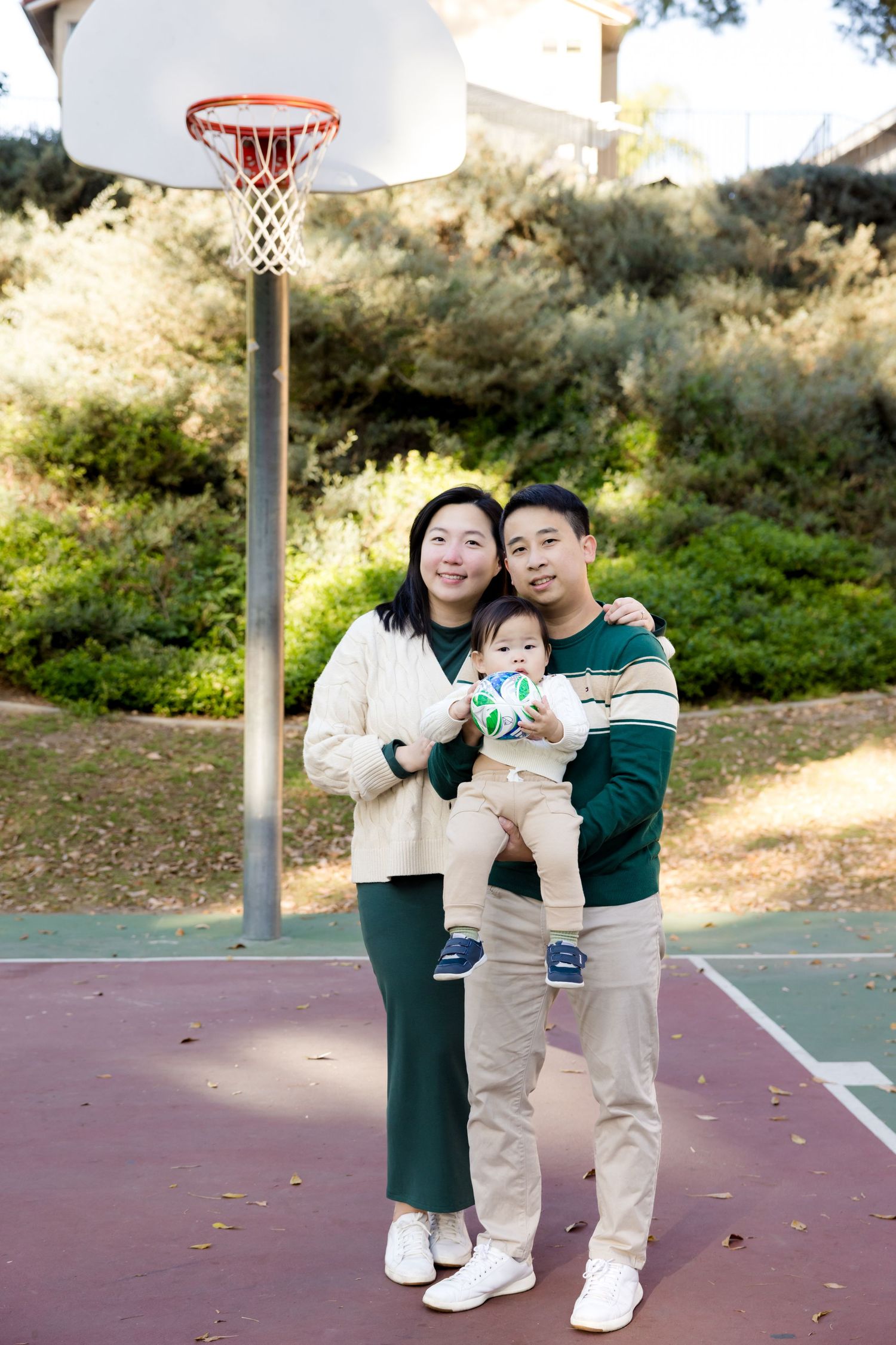 A couple with a baby take photos on an outdoor basketball court with a hoop in the background.