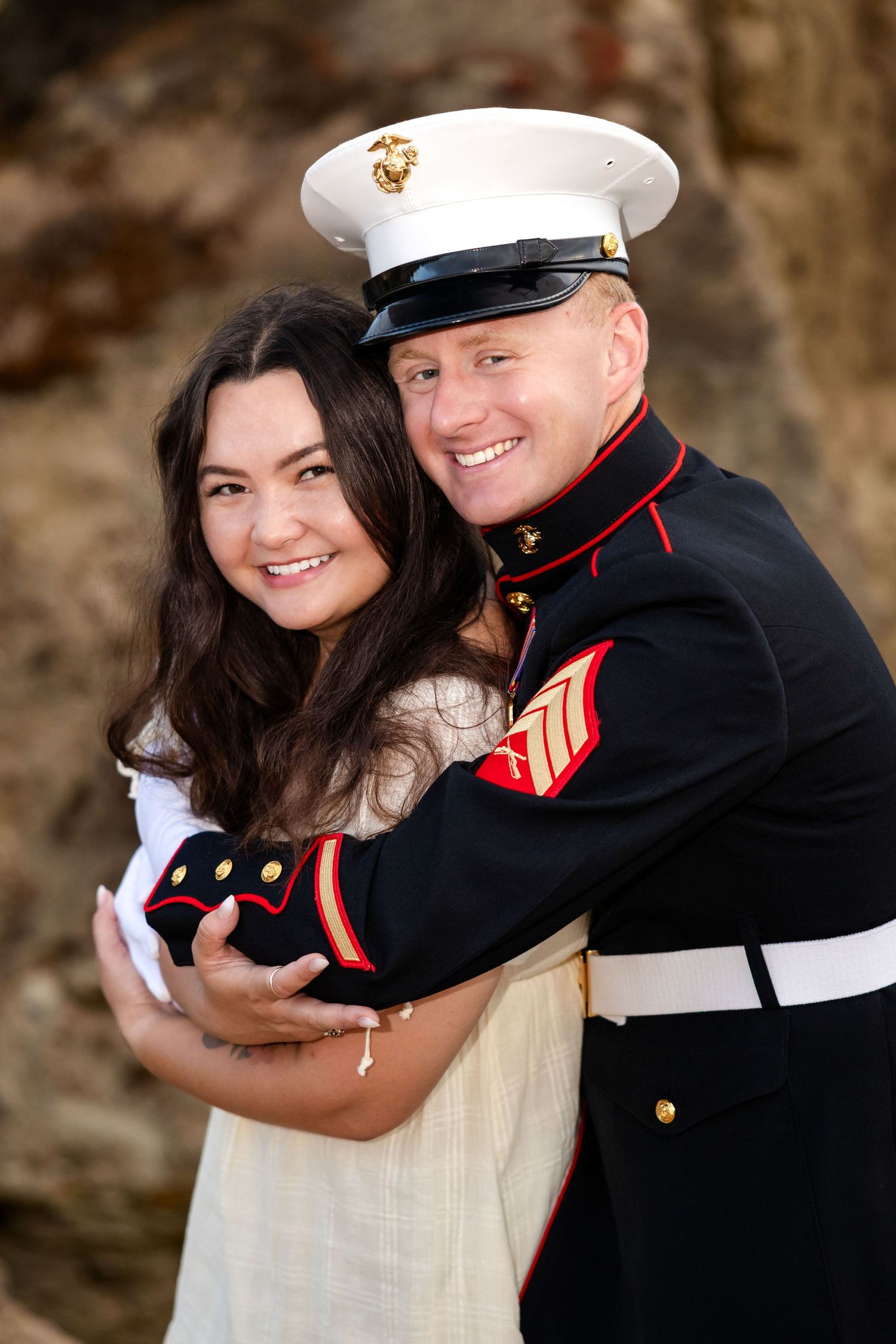 US Marine in dress blues uniform embracing smiling woman outdoors, couple portrait photo.