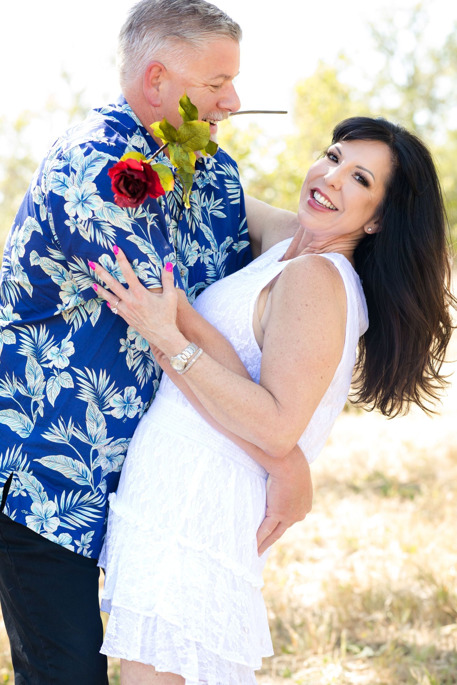 A couple in white and blue floral attire share a romantic moment outdoors with a red rose boutonniere.