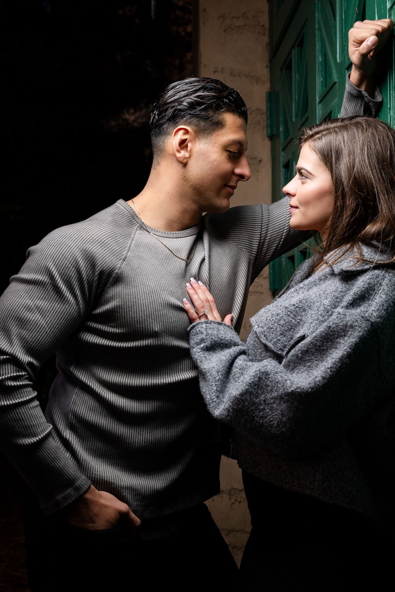 A couple in dark sweaters share an intimate dance-like embrace against a shadowy background.