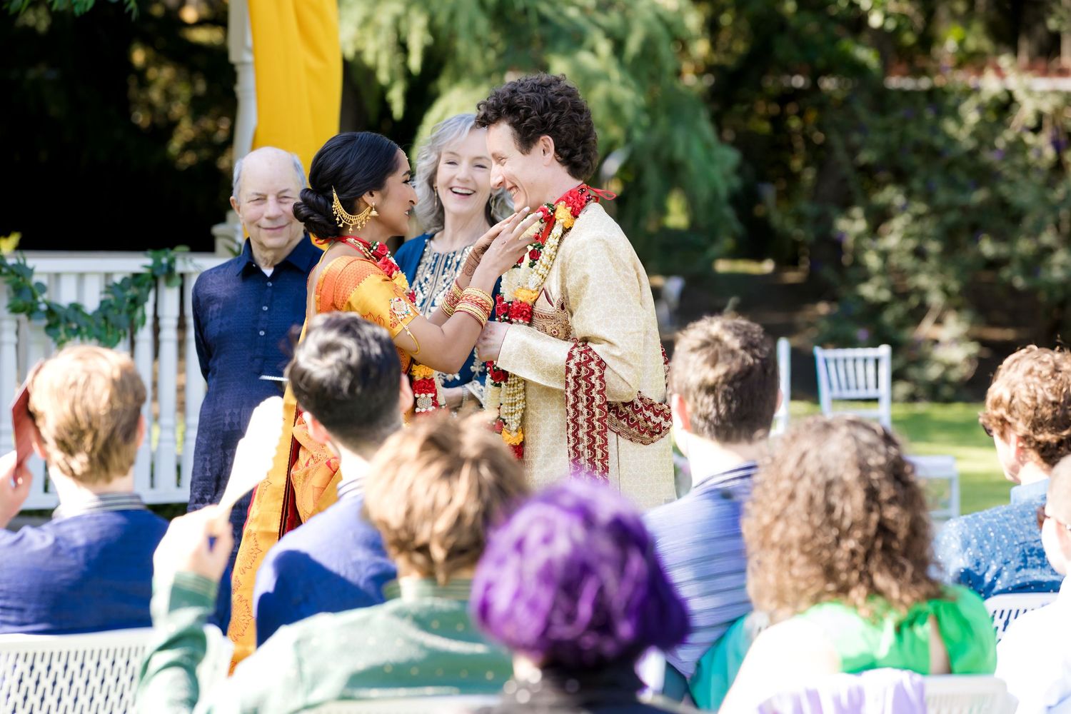 A wedding ceremony takes place outdoors with guests seated in white chairs watching a couple at the front.