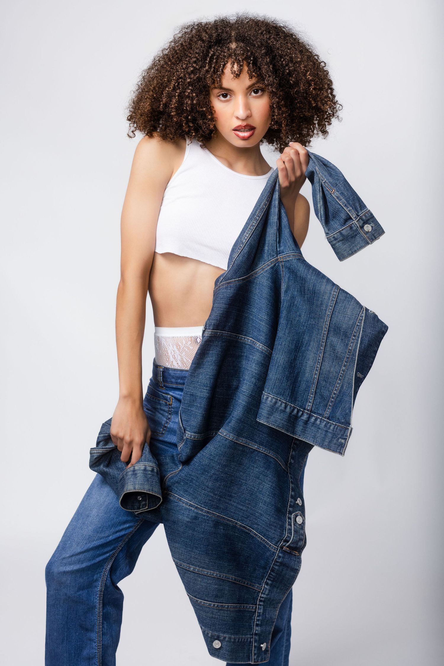 Fashion model wearing white crop top and oversized denim jeans poses against light background in studio photoshoot.