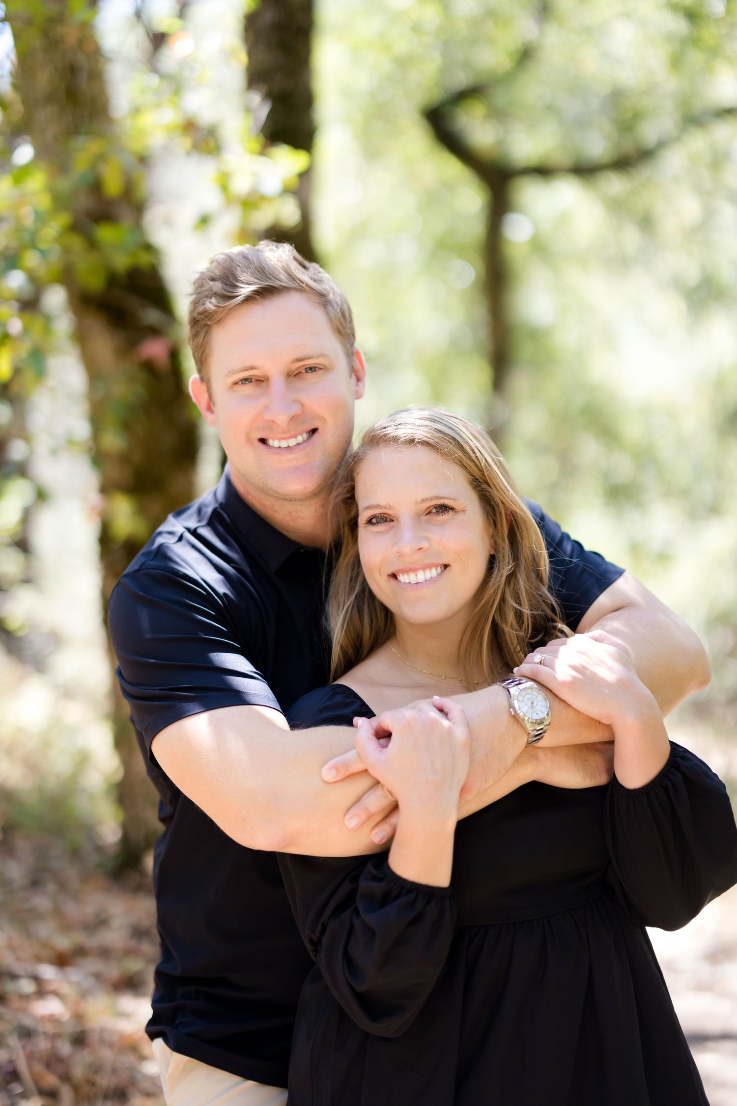 A couple in matching black outfits share tender embraces while posing outdoors in a sunlit wooded setting.