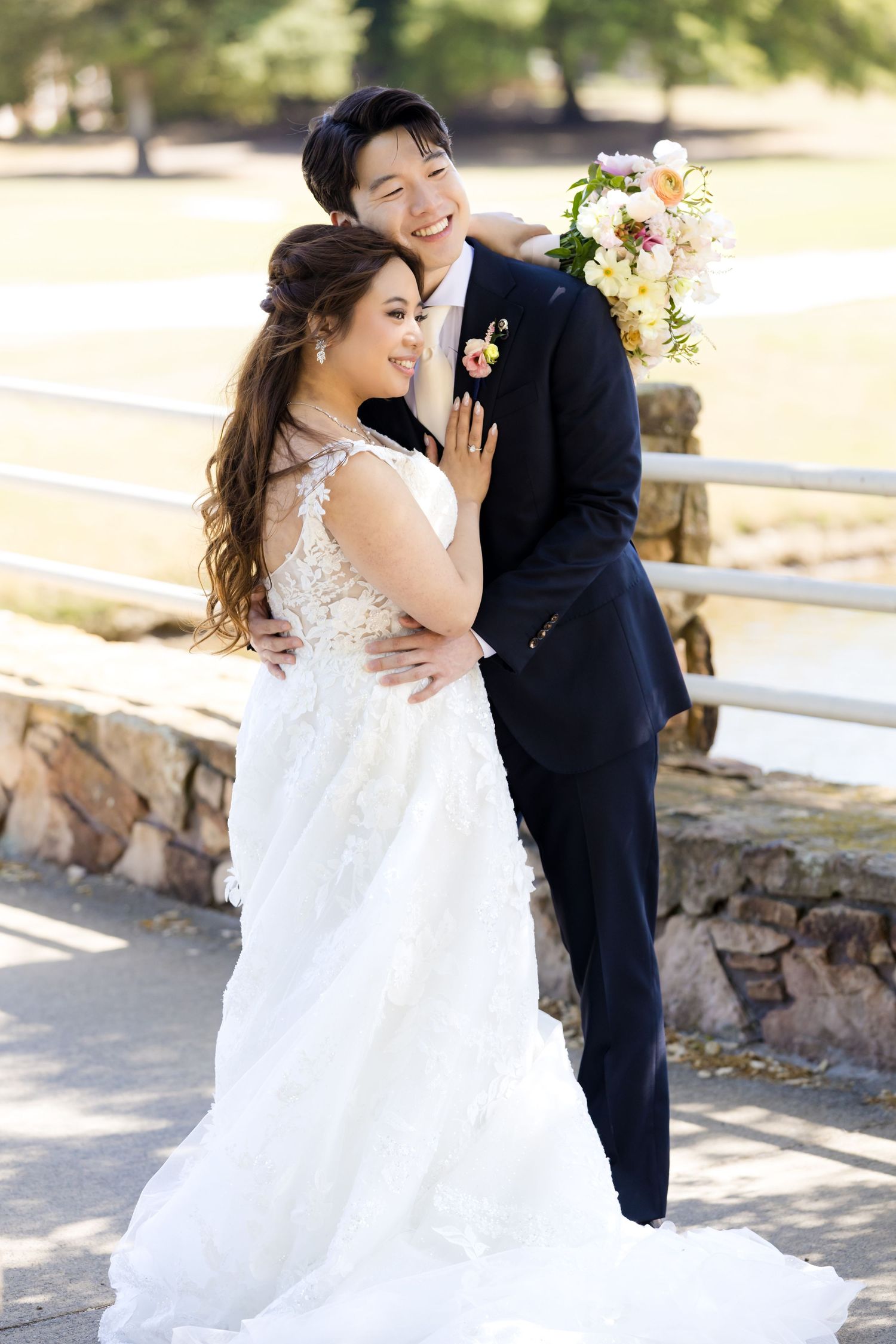 A couple in wedding attire embrace outdoors on a sunny day, with the bride in a lace white gown and groom in navy suit.