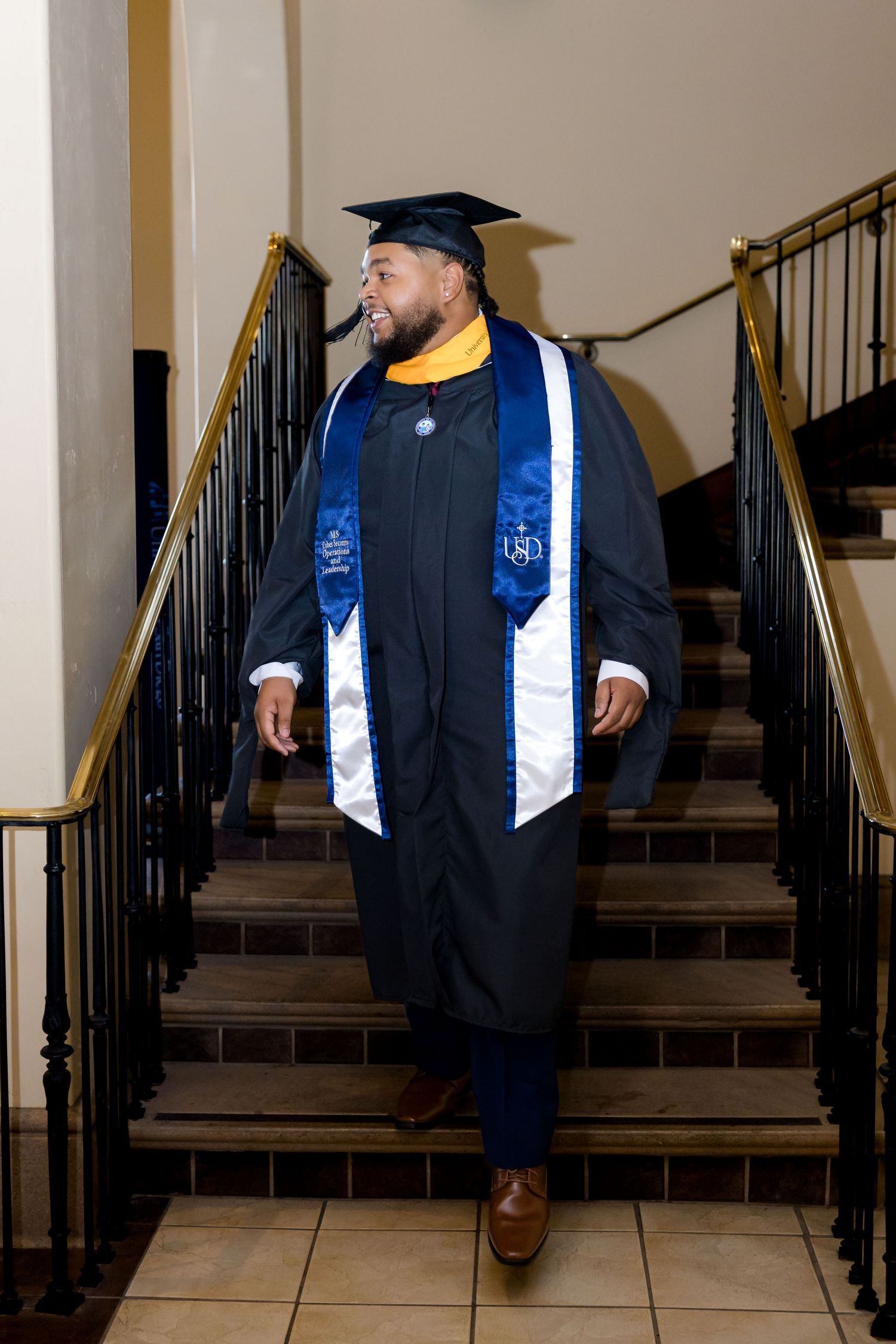 A man in black graduation gown, cap, and blue sash walks down stairs during his commencement ceremony.