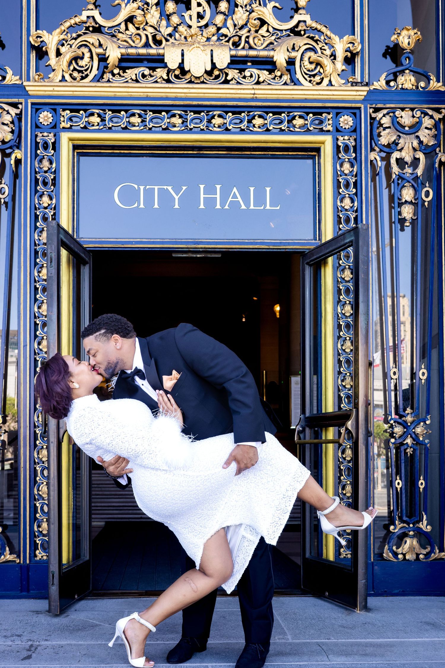 A romantic couple in formal attire poses for wedding photos in front of ornate blue and gold City Hall doors.