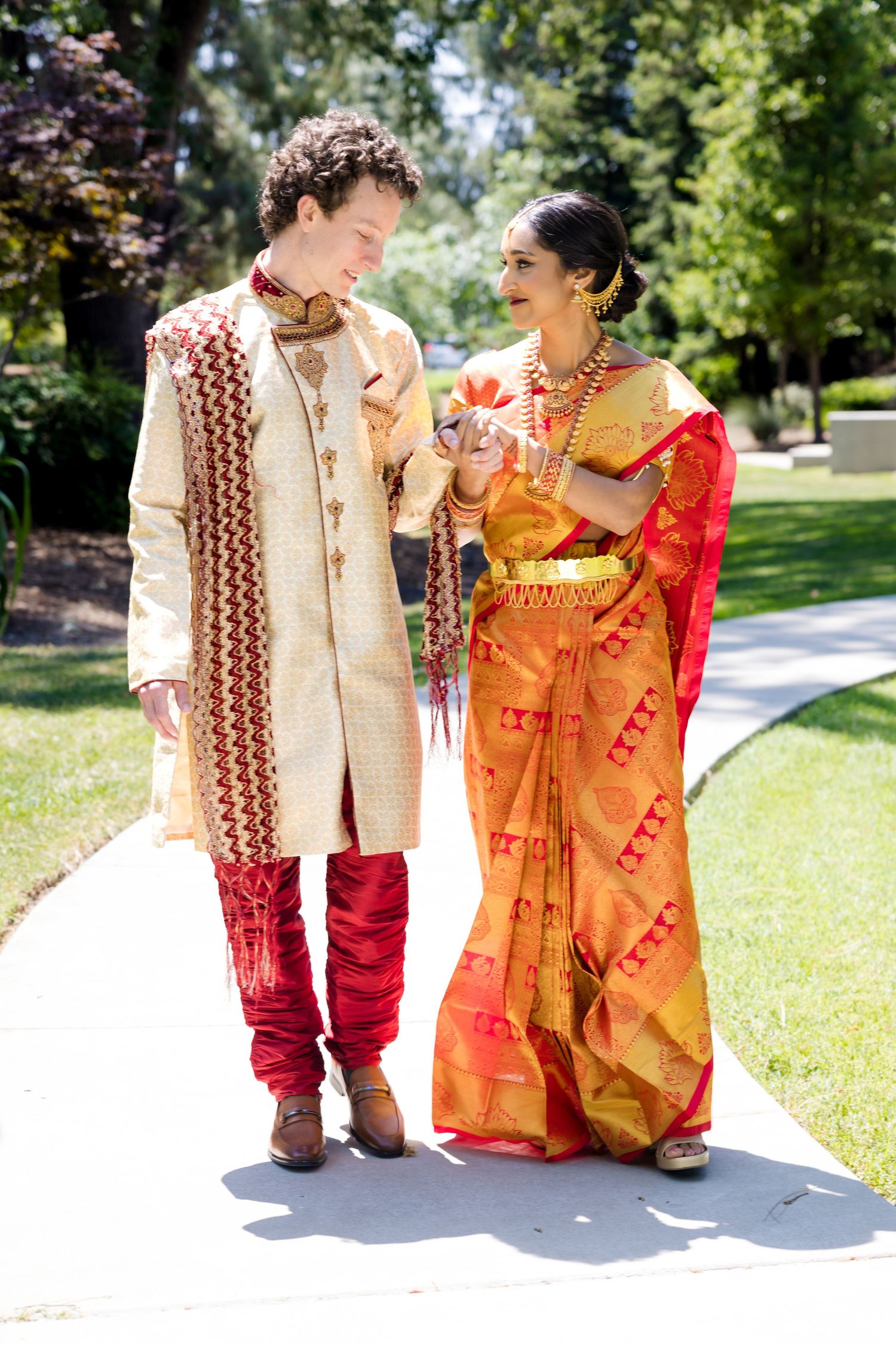 Couple in traditional Indian wedding attire walking together on sunny garden path.