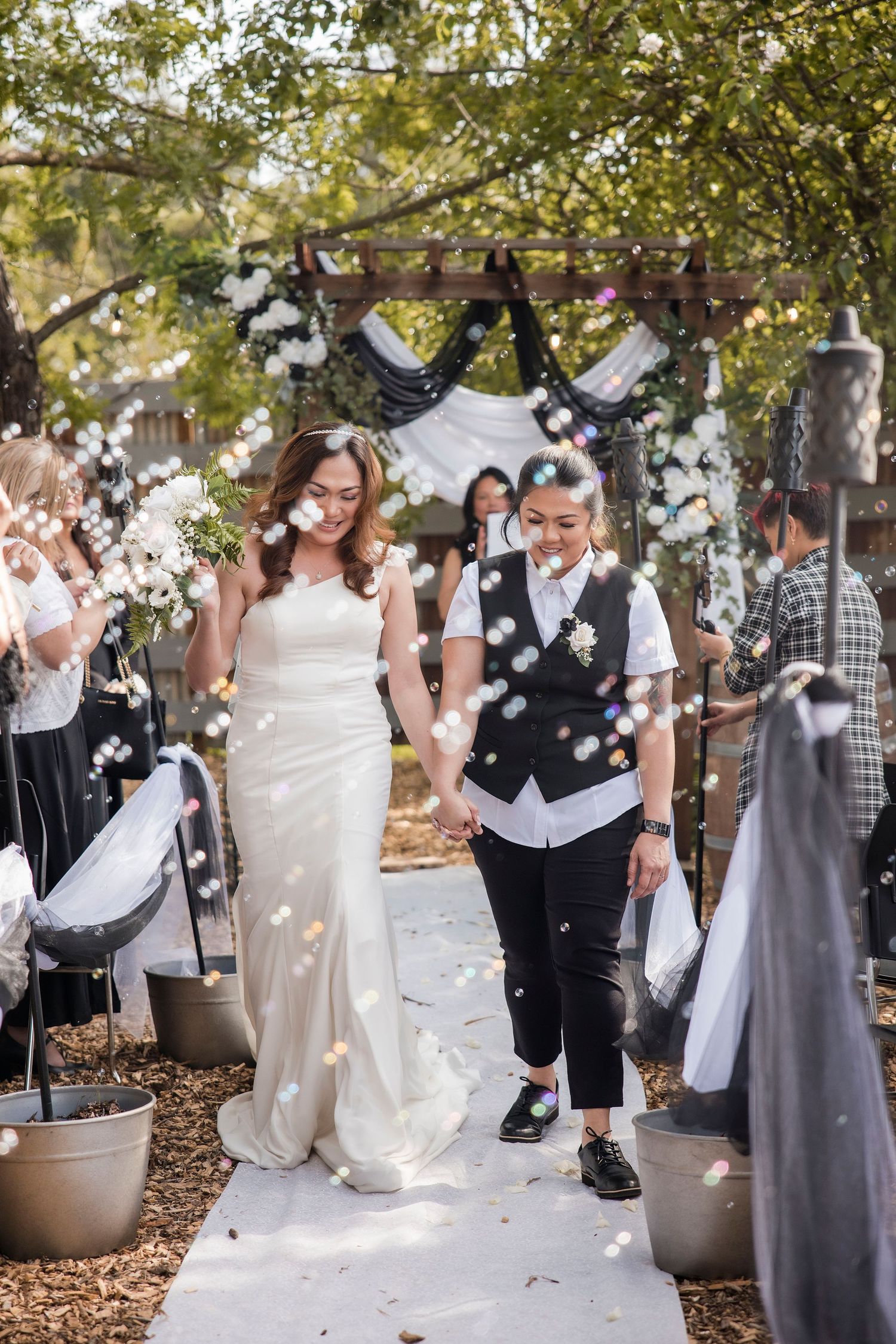 Wedding couple walking down decorated outdoor aisle while guests throw confetti.
