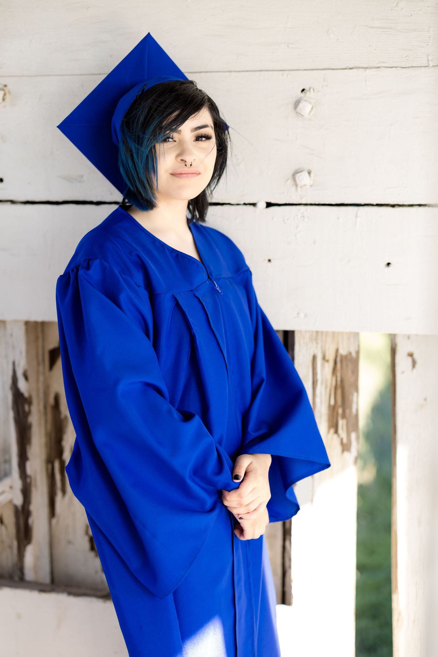 Graduate in royal blue cap and gown poses against white wooden wall.