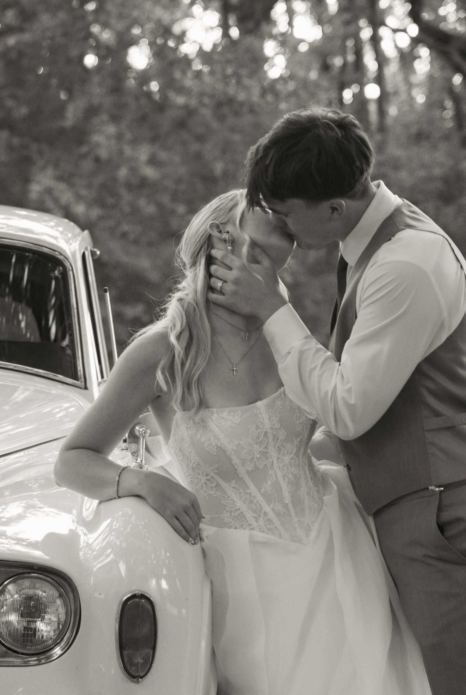 Black and white wedding photo of bride and groom sharing a tender kiss beside a vintage car outdoors.